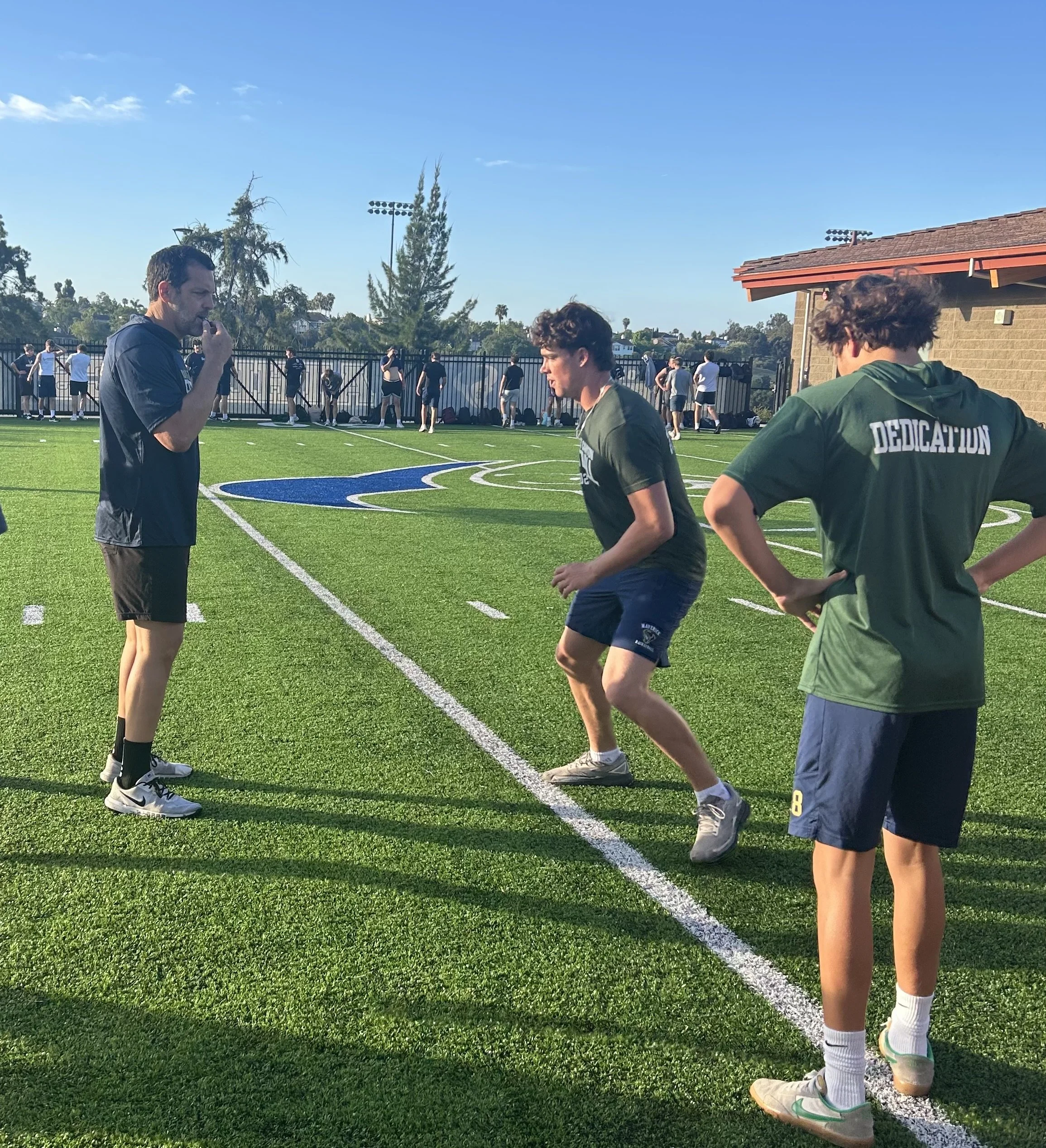 Group of young men on a sports field, some standing and some walking, with a coach or instructor speaking to one of them. The field has bright green grass, marked lines, and a blue and white logo. In the background, there are trees, a fence, and a small building with a red-tiled roof under a clear blue sky.
