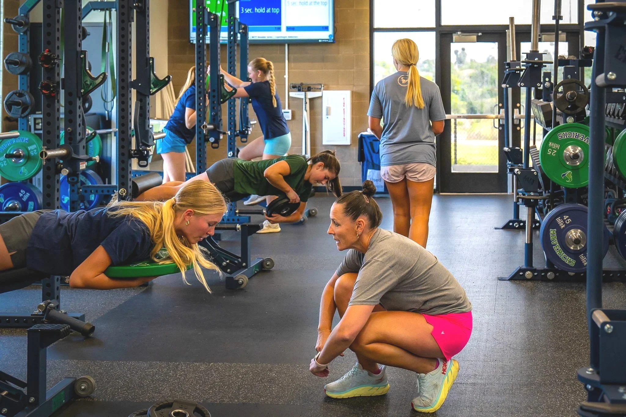 A gym scene with multiple women performing various exercises on the floor and weight machines. One woman in pink shorts and white sneakers is stretching or doing a floor exercise, while others are lifting weights or holding workout pads. The gym has weight racks, a large window, and a TV screen in the background.