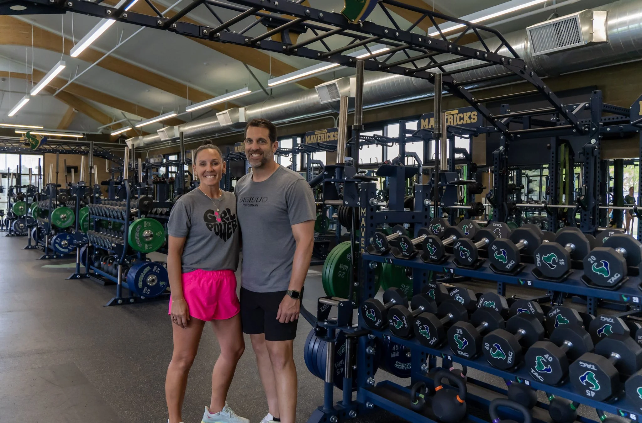 A man and a woman smiling and standing together in a gym. The gym has various weights and exercise equipment in the background, with large windows and overhead lighting.