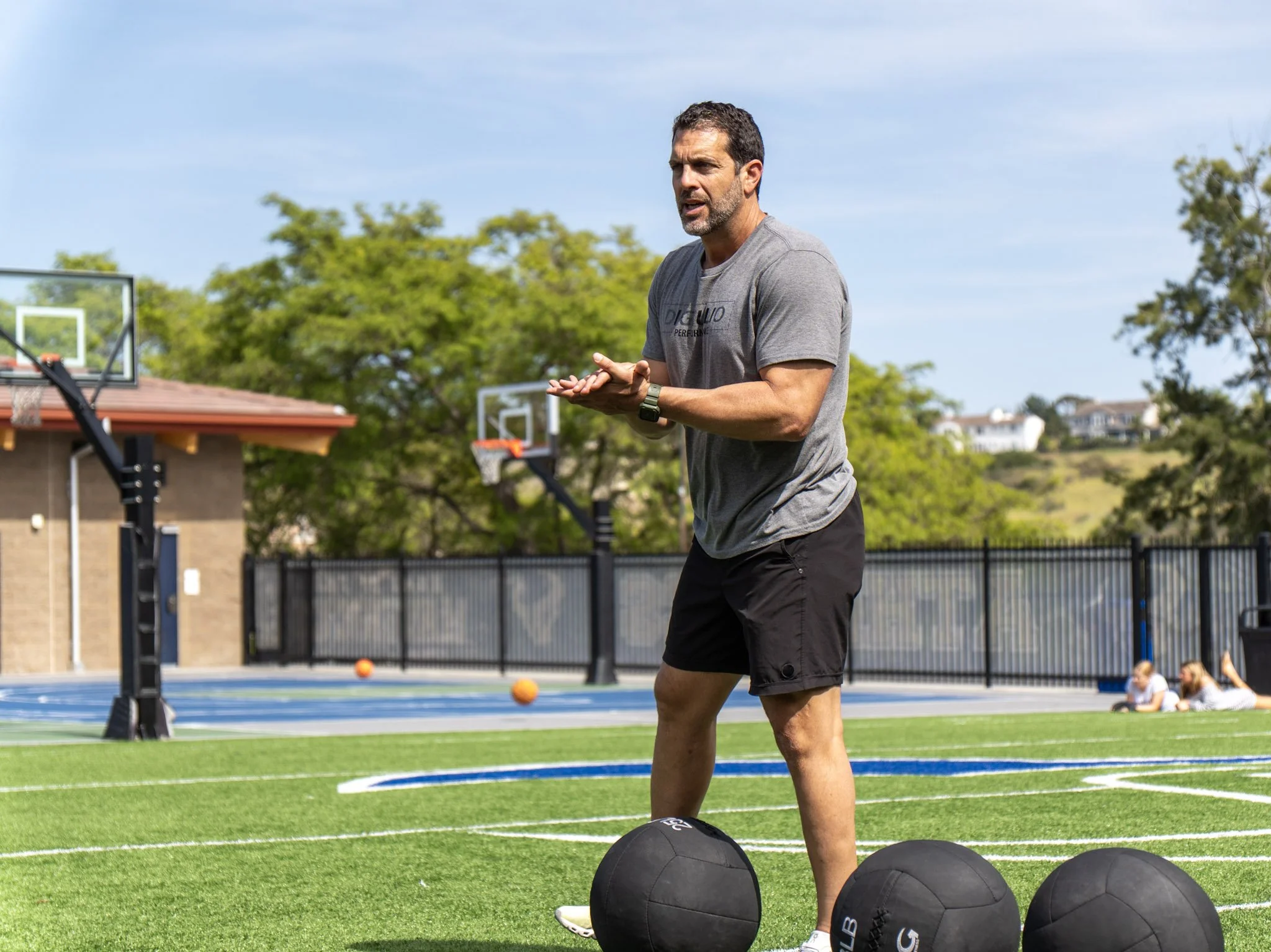 A man standing on a sports field near basketball hoops, wearing a gray t-shirt and black shorts, with large black exercise balls in front of him and several children sitting on the ground in the background.