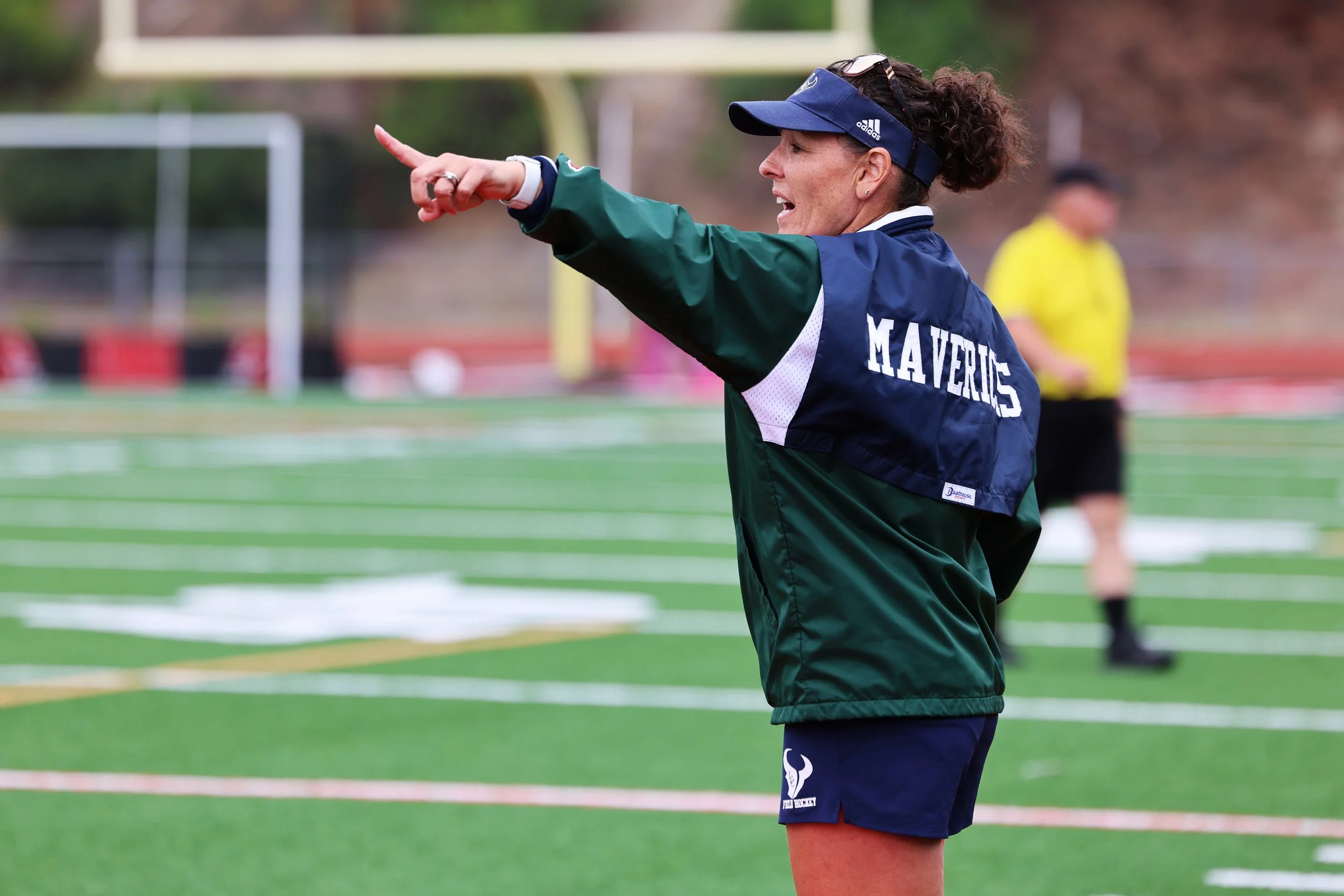 A woman wearing a navy visor and green jacket, pointing and speaking on a sports field during a sporting event.