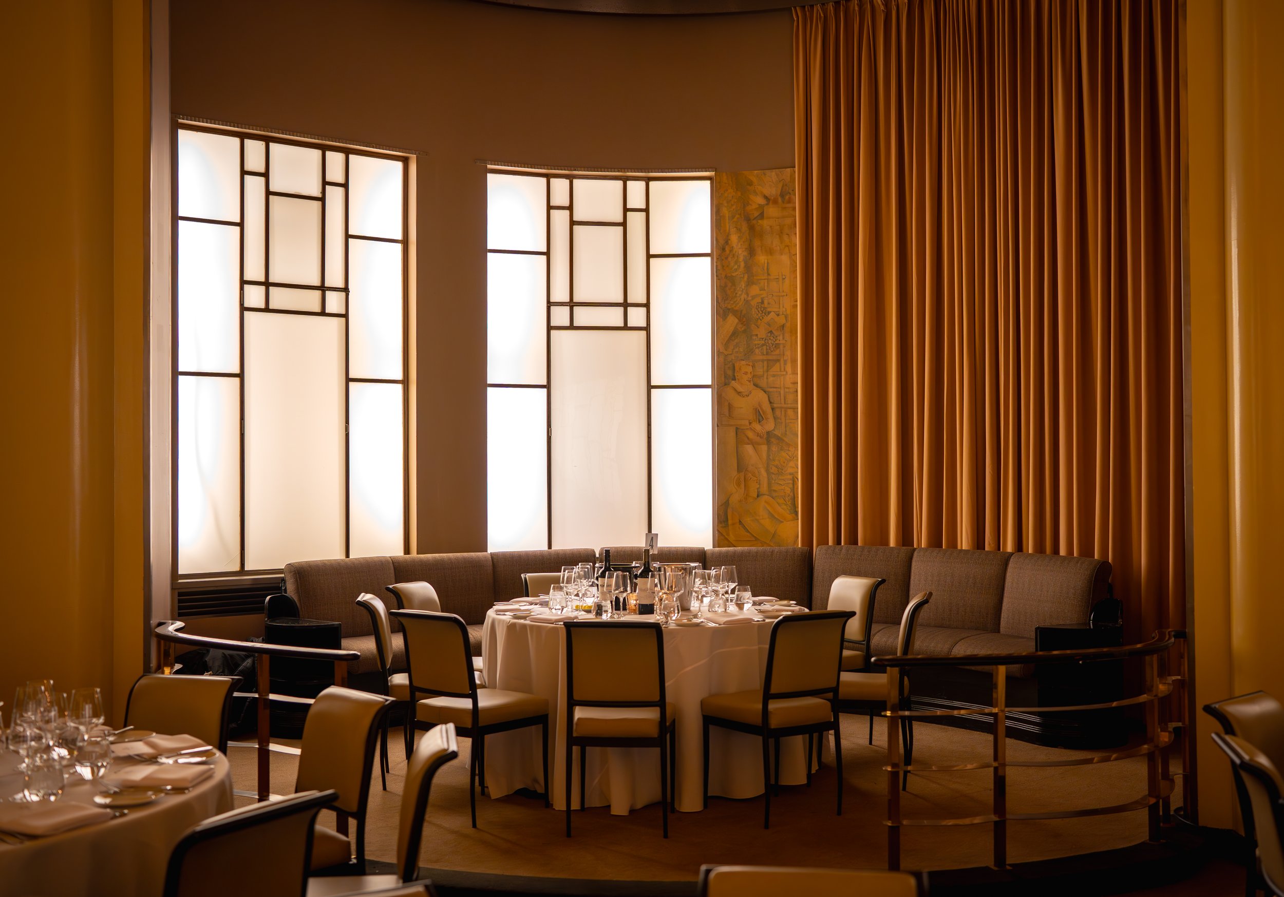 Elegant restaurant interior with round tables, white tablecloths, set for dining. Modern art deco windows, beige chairs, and gold curtains create a warm ambiance.