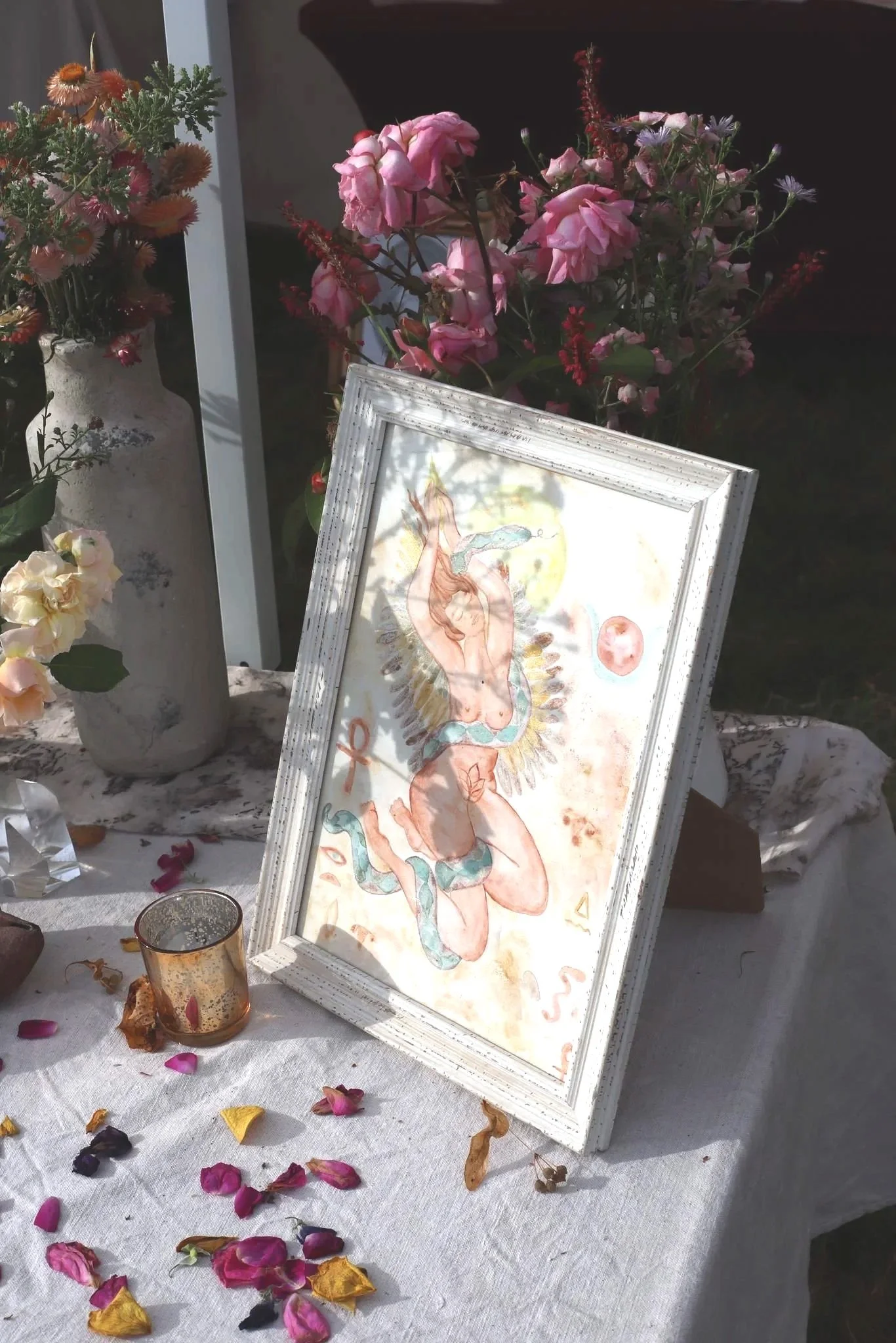 A framed artwork featuring a nude woman with a snake around her body, decorated with feathers and more celestial elements, placed on a table with flower petals and decorative items, surrounded by vases of pink and cream flowers.