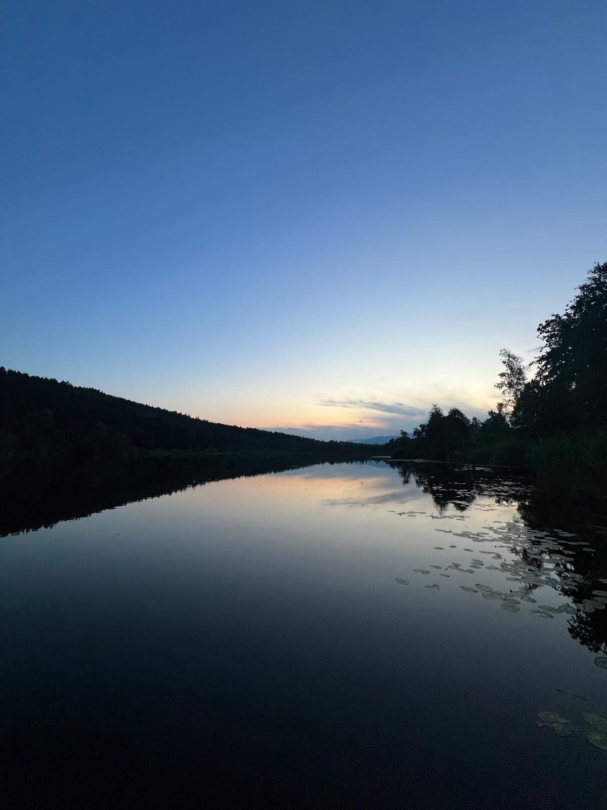 Ein ruhiger See bei Sonnenuntergang, umgeben von Bäumen und Hügeln, mit spiegelnder Wasseroberfläche