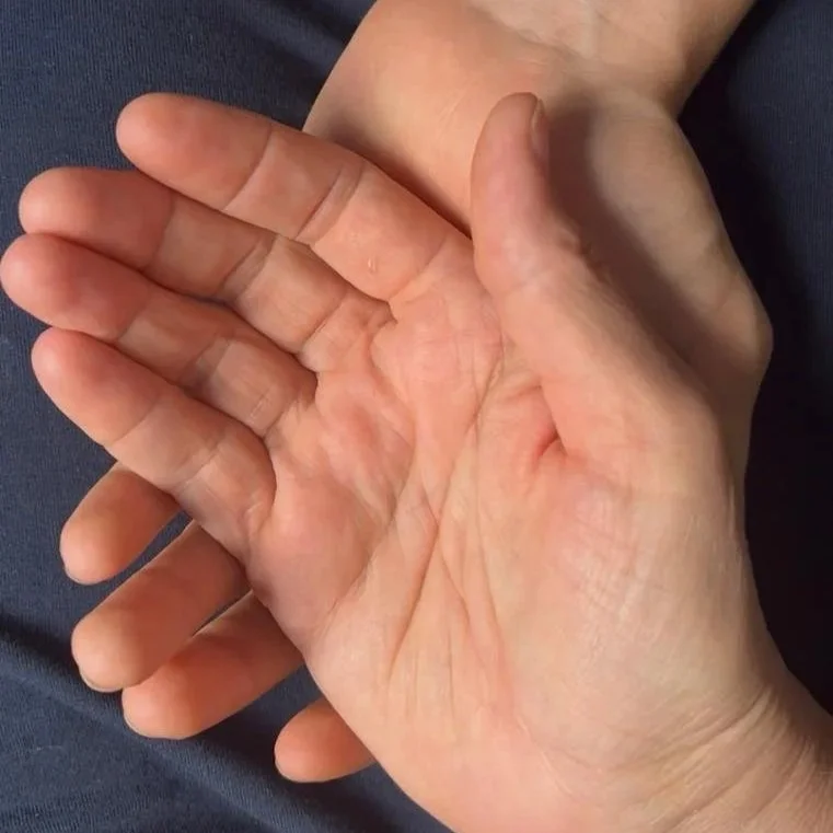 Zwei Hände liegen sanft ineinander, die eine Hand trägt die andere, auf einem dunklen Stoffhintergrund.