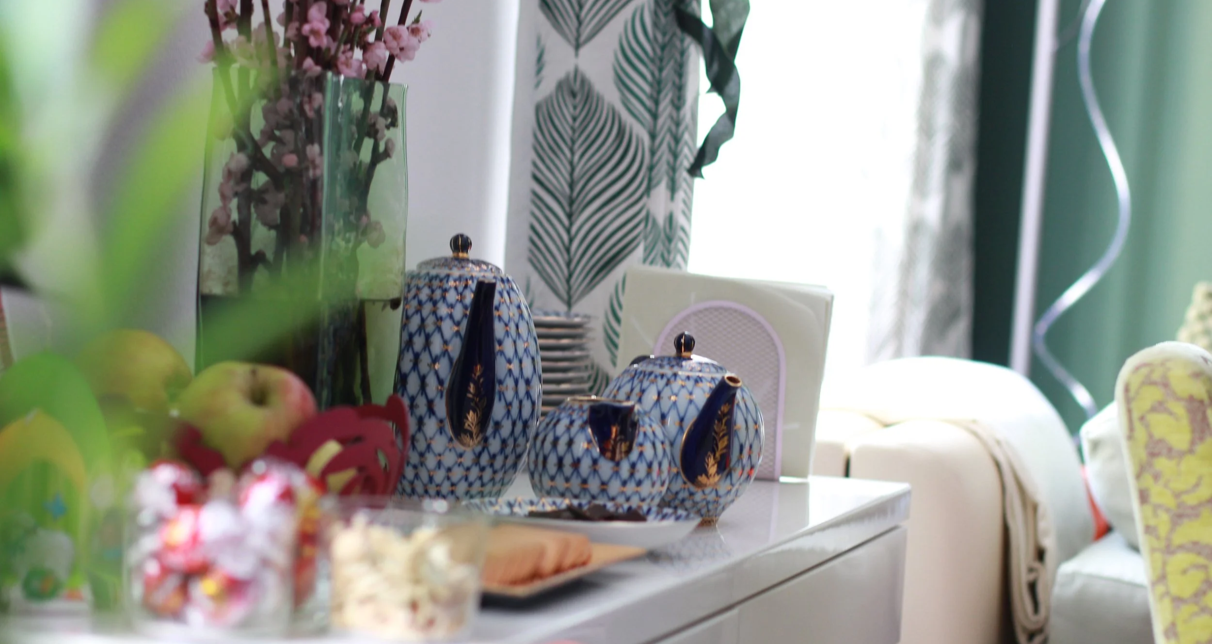 Bedeckter Tisch mit blauen Teekannen, Obst und Blumen, im Hintergrund Gardinen mit Pflanzenmuster