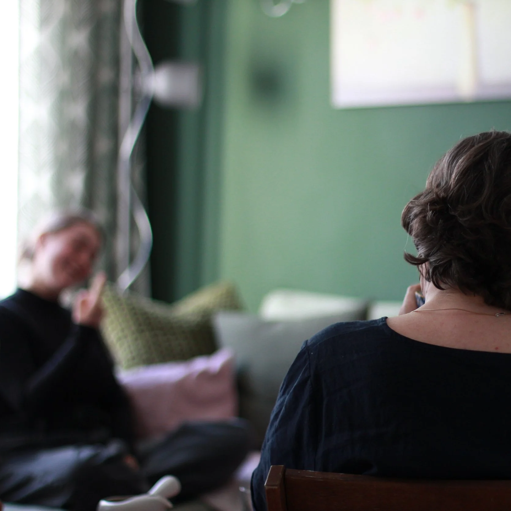 Menschen sitzen in einem Raum, eine Person im Vordergrund mit kurzen braunen Haaren, eine weitere Person links mit längeren blonden Haaren, alle an Tischen, im Hintergrund grüne Wände und Fenster.