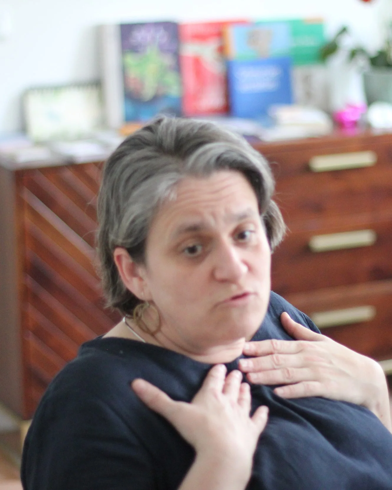 Eine Frau mit kurzen grauen Haaren, die ein dunkelblaues Oberteil trägt, zeigt mit beiden Händen an ihre Brust, während sie in die Kamera schaut. Im Hintergrund sind bunte Bücherregale und eine grüne Pflanze sichtbar.