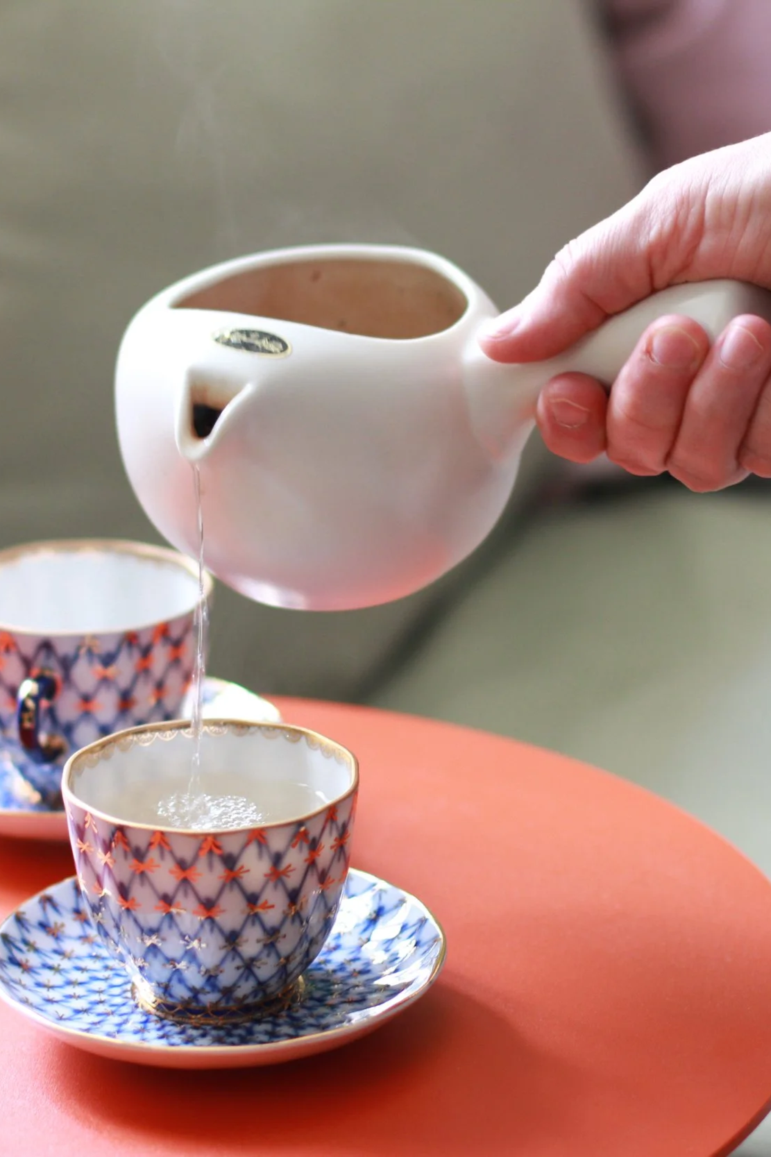 Person gießt Tee aus einer weißen Teekanne in eine Teetasse mit goldenen Akzenten auf einem orangefarbenen Tisch.