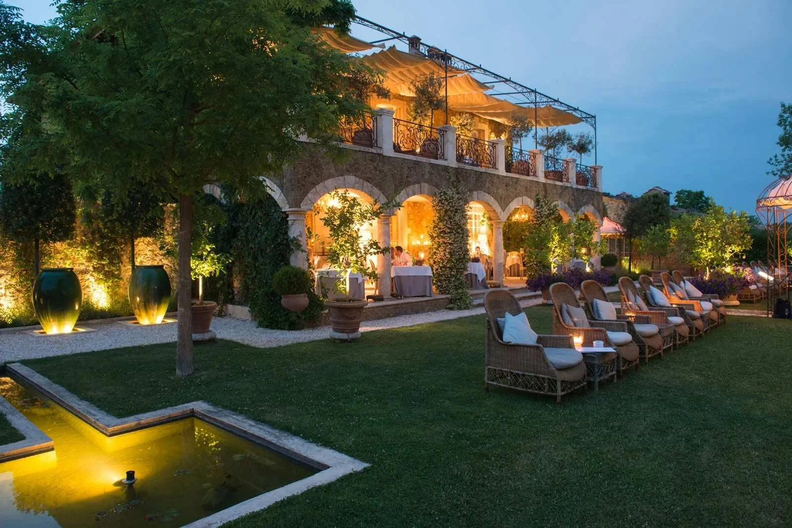 Borgo Santo Pietro in Tuscany at dusk, a stone arched loggia lit from within with wicker loungers on the lawn beside a reflecting pool.