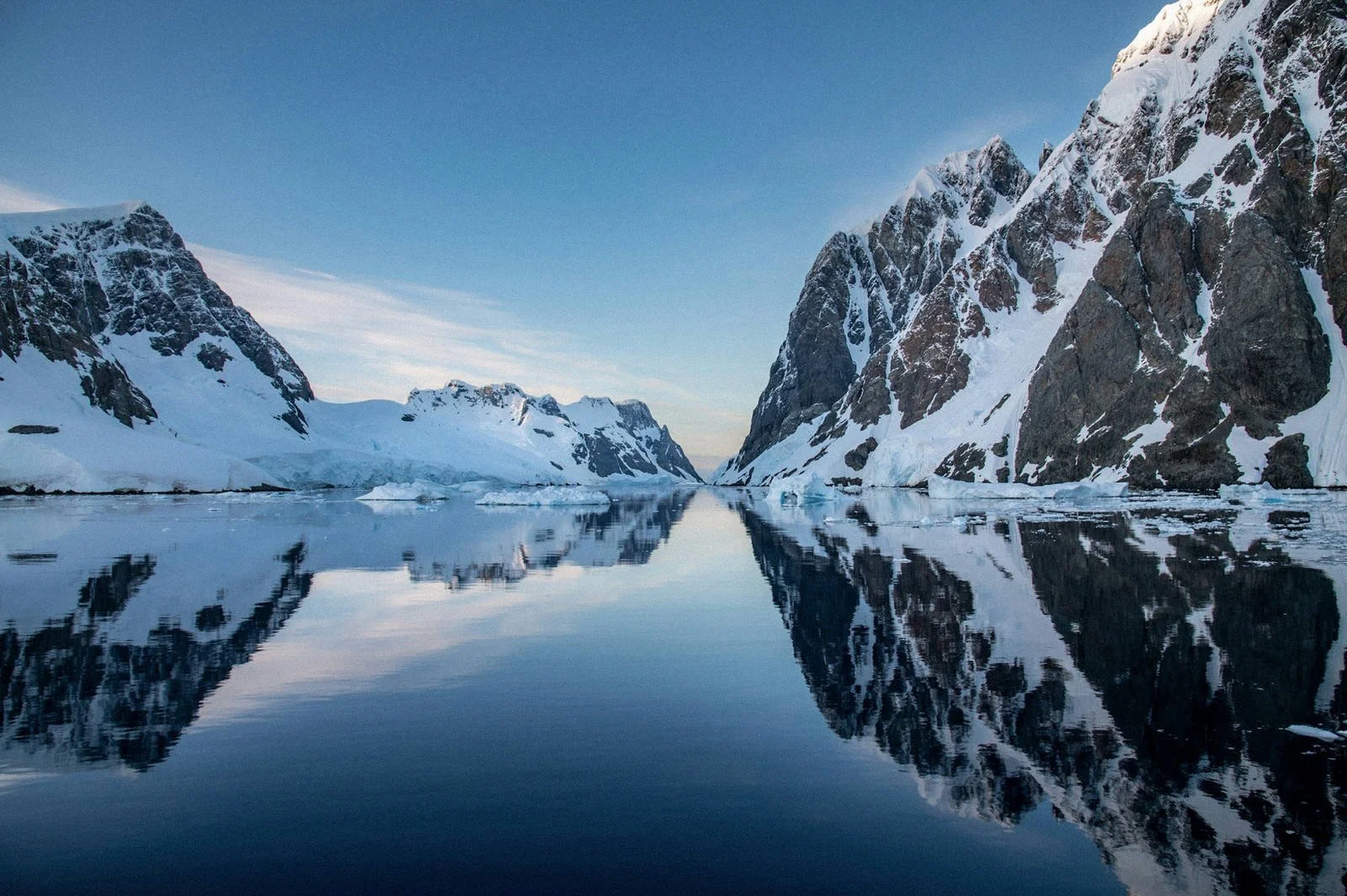 Steep dark rock faces with patchy snow reflected in calm water in a narrow Antarctic channel late in the season.