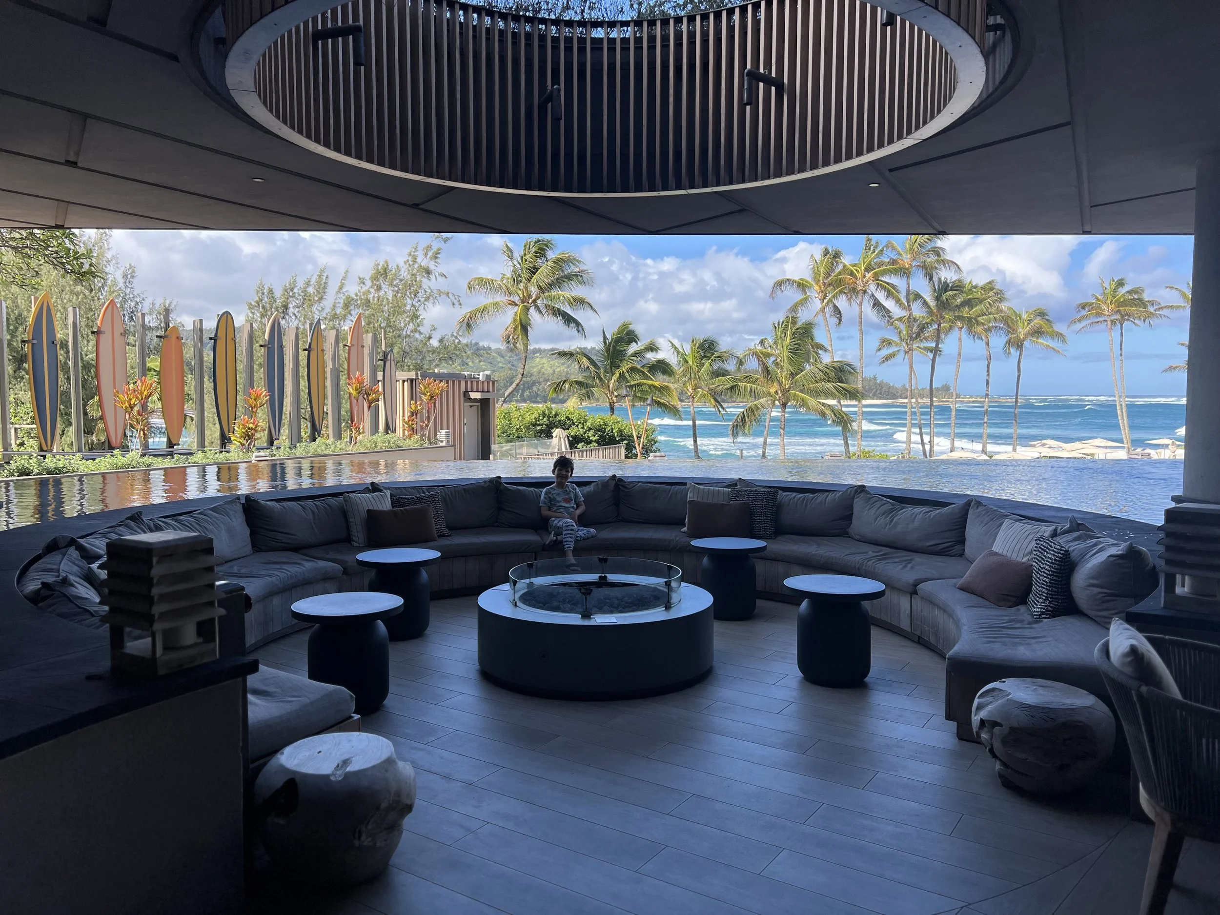 Kate Van Dell’s son seated in the oceanfront lobby at The Ritz-Carlton O‘ahu, Turtle Bay, with surfboards, palm trees and an ocean view