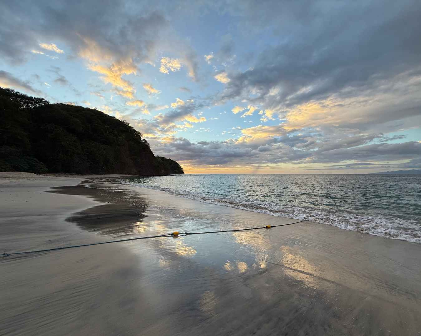 Sunset over a beach with cloudy sky, gentle waves, and a wooded hill on the left; the shoreline has a rope with yellow floats.