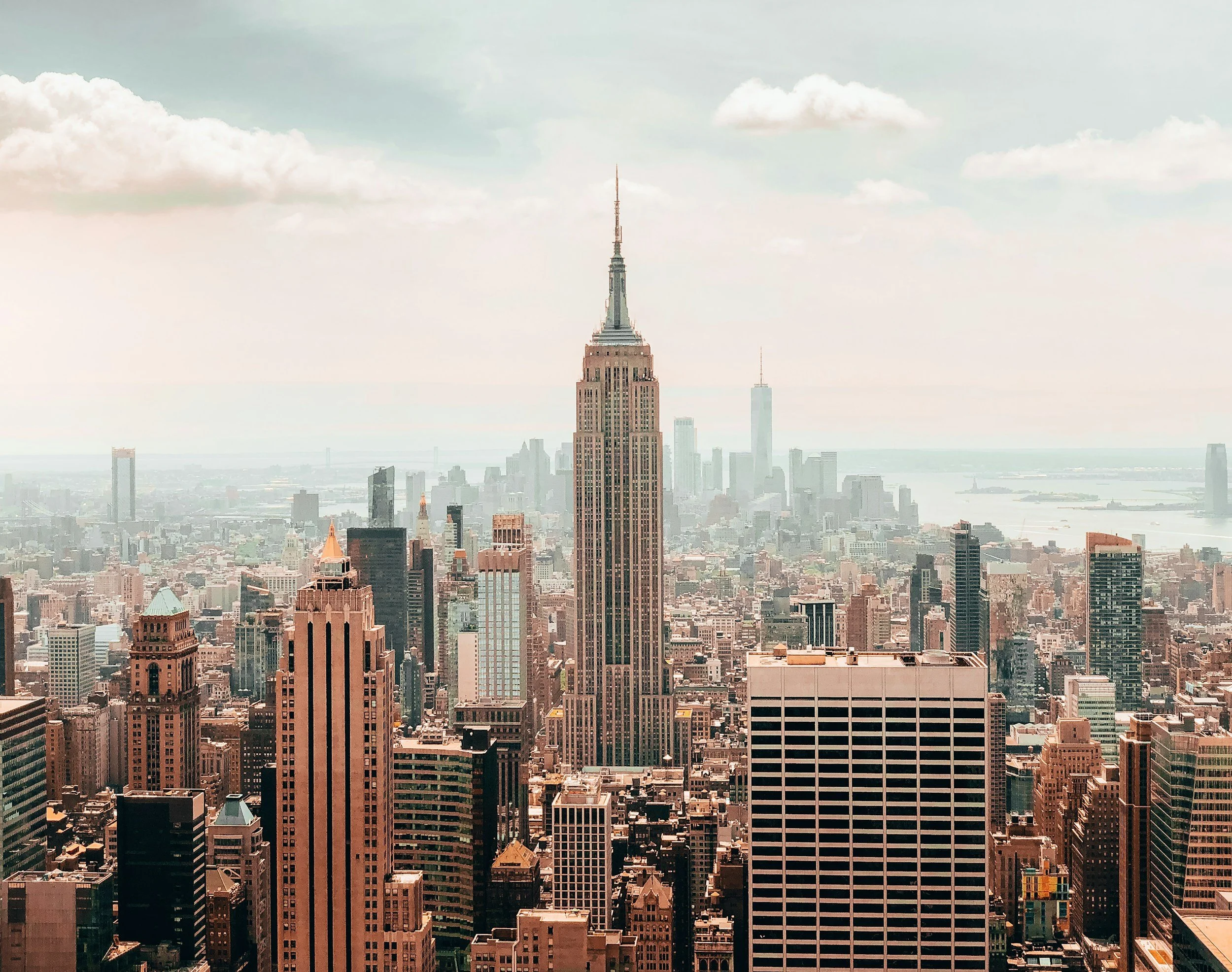 A panoramic cityscape of New York City featuring the Empire State Building in the center, surrounded by numerous skyscrapers, with the Hudson River and distant buildings in the background under a partly cloudy sky.