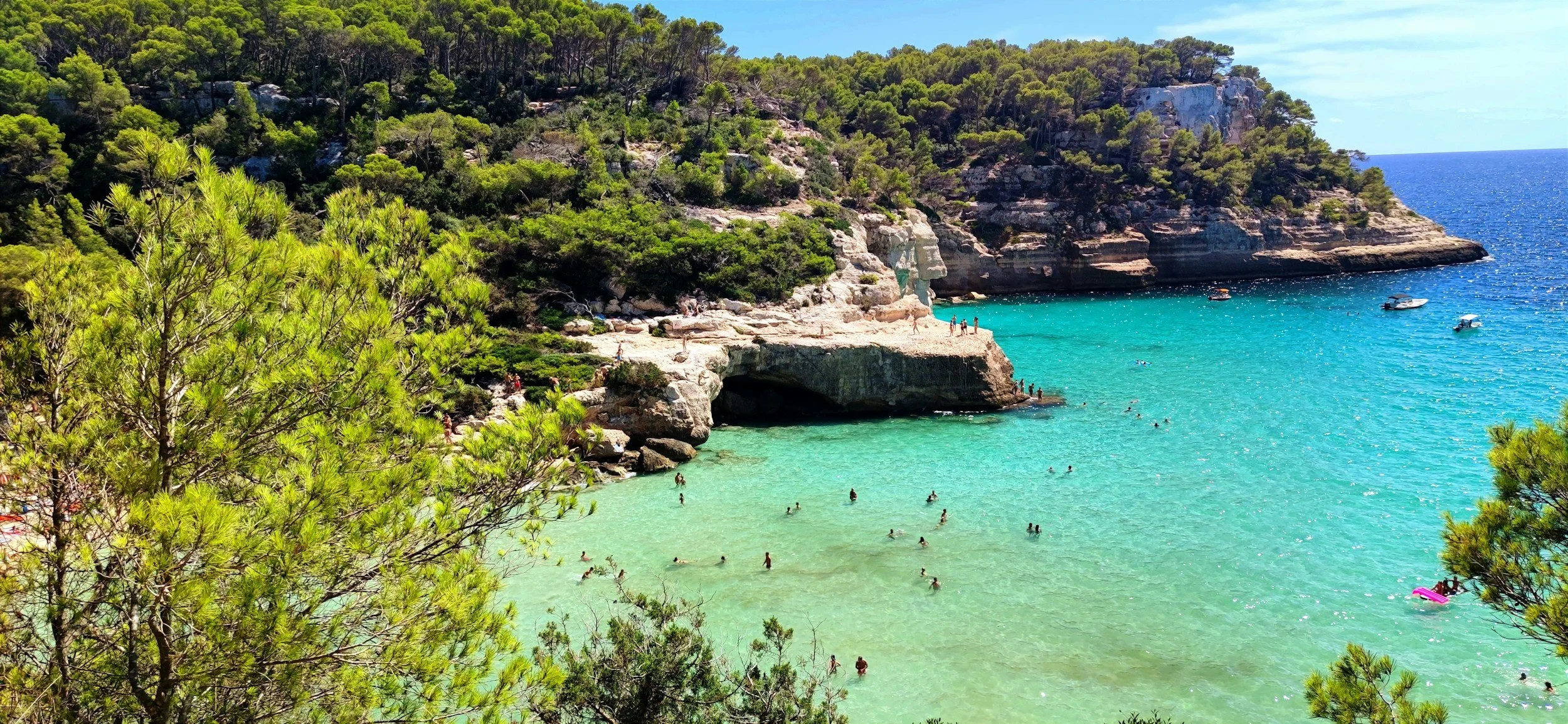 Swimmers in clear turquoise water beneath pine-covered limestone cliffs at Cala Mitjana beach in Menorca.