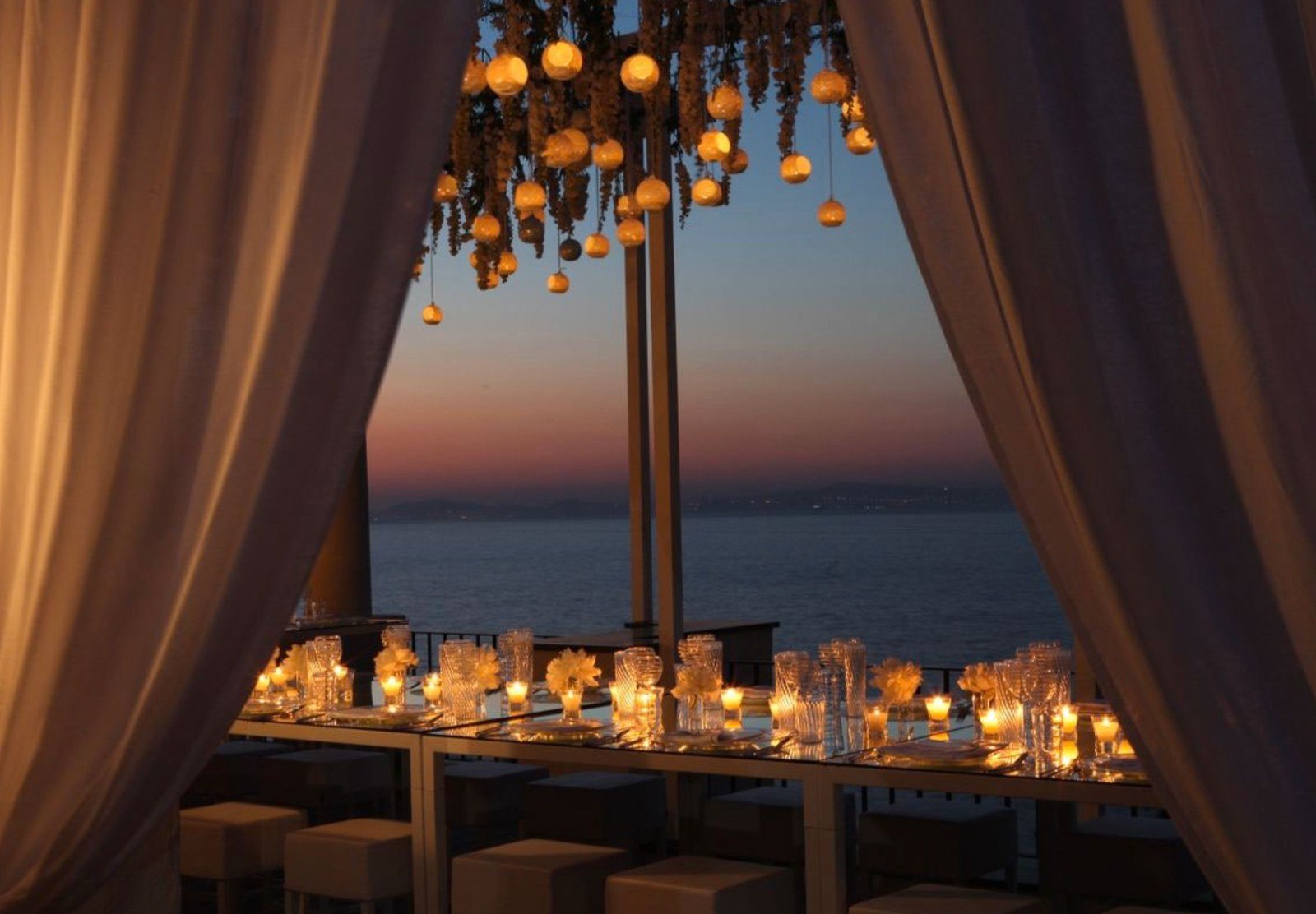 Candlelit evening dinner at J.K. Place Capri, draped curtains framing a sea view at sunset with a hanging candle installation above a mirrored table.
