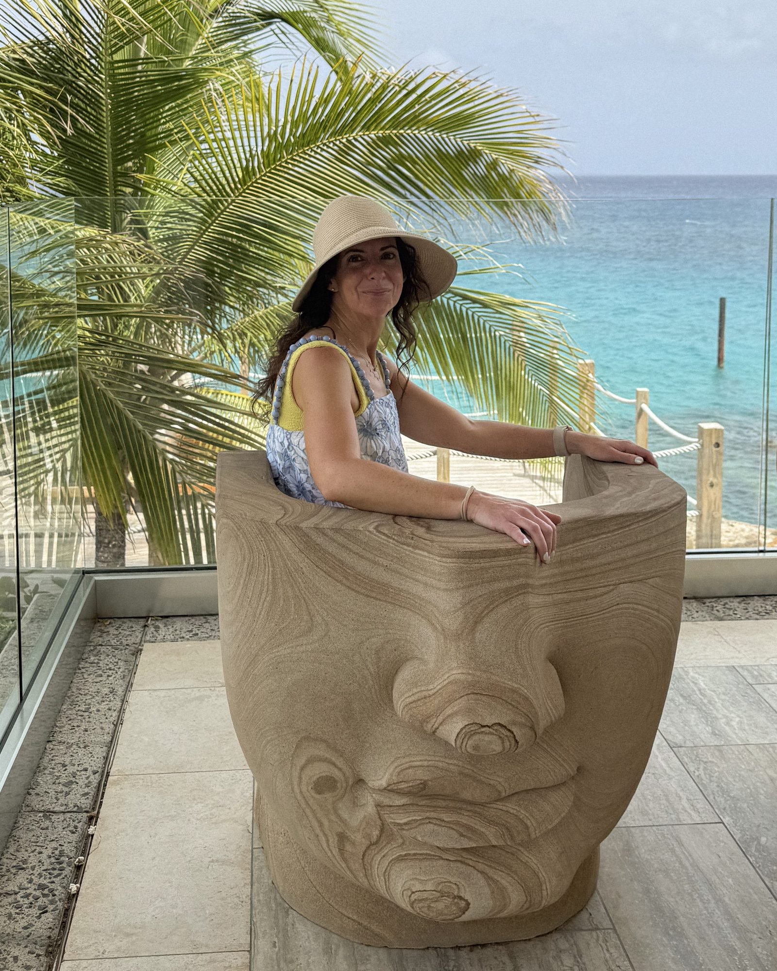 Luxury travel advisor, Kate Van Dell, sitting in the iconic face chair at Four seasons anguilla with turquoise water and palm tree in the background