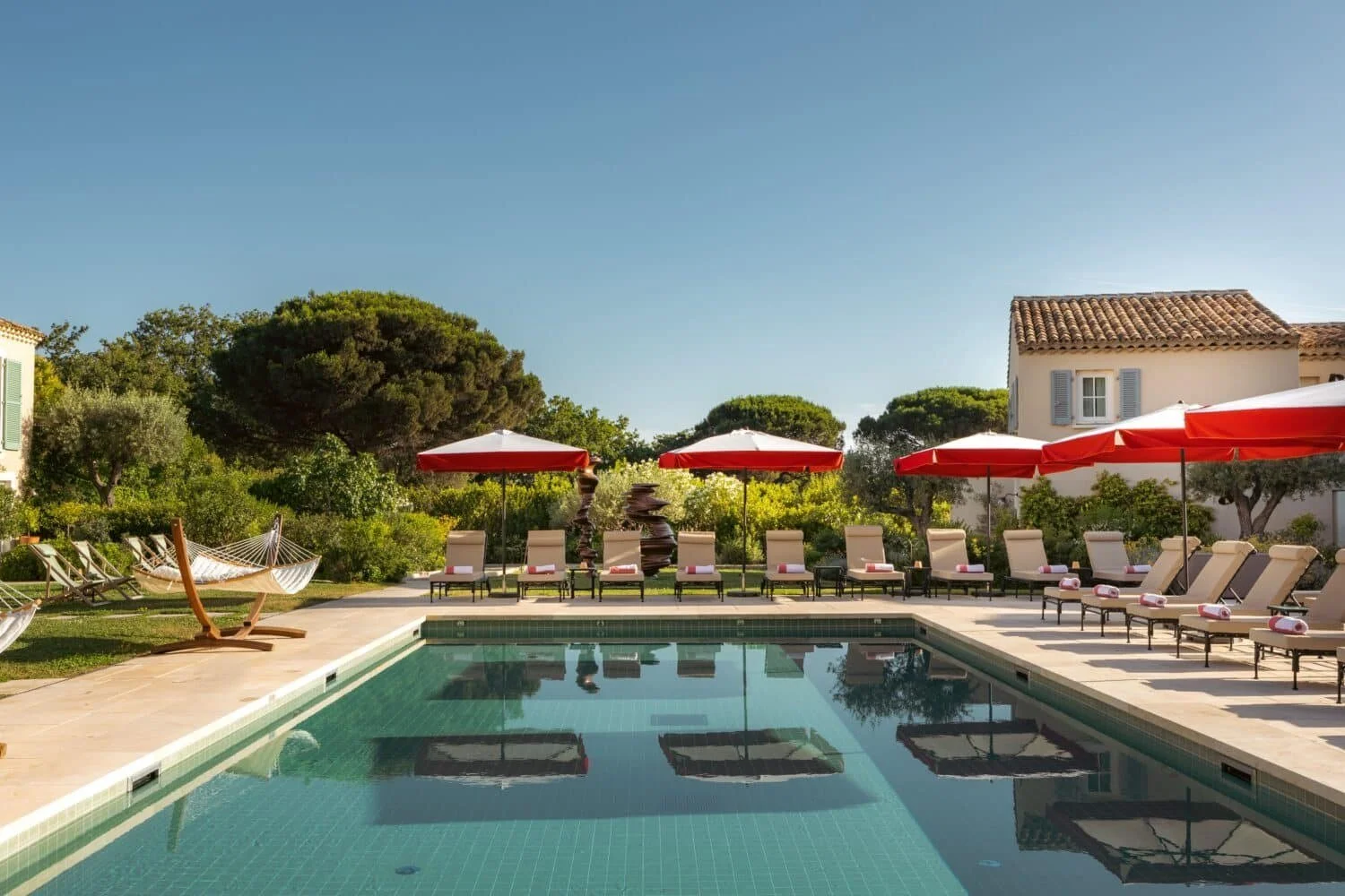 Swimming pool at Lou Pinet in St-Tropez, red-and-white parasols and loungers beside a Provencal house with blue shutters and a hammock under the pines.