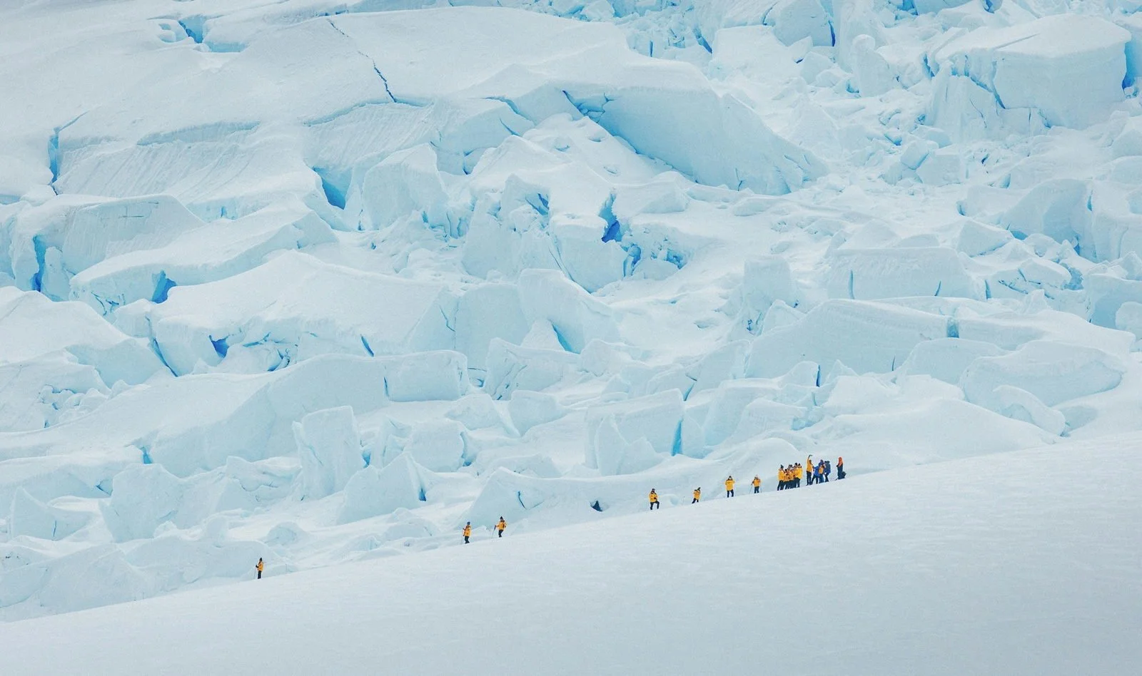 Expedition passengers in yellow parkas walking single file across deep snow beneath a large crevassed glacier on the Antarctic Peninsula.