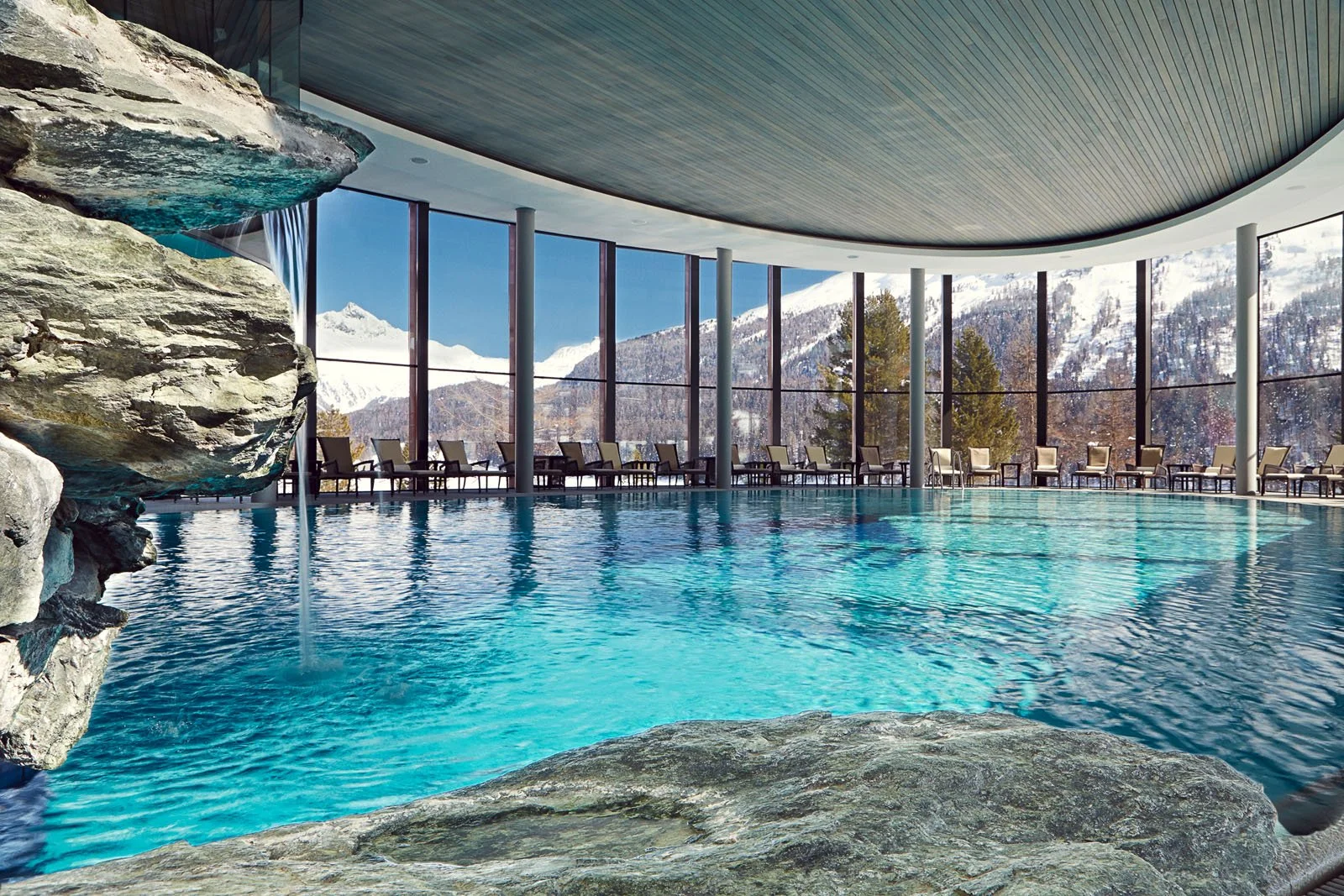 Indoor swimming pool with large windows showing snow-capped mountains and trees outside. Rock formation with a waterfall feature on the left, surrounded by lounge chairs along the windowed wall.