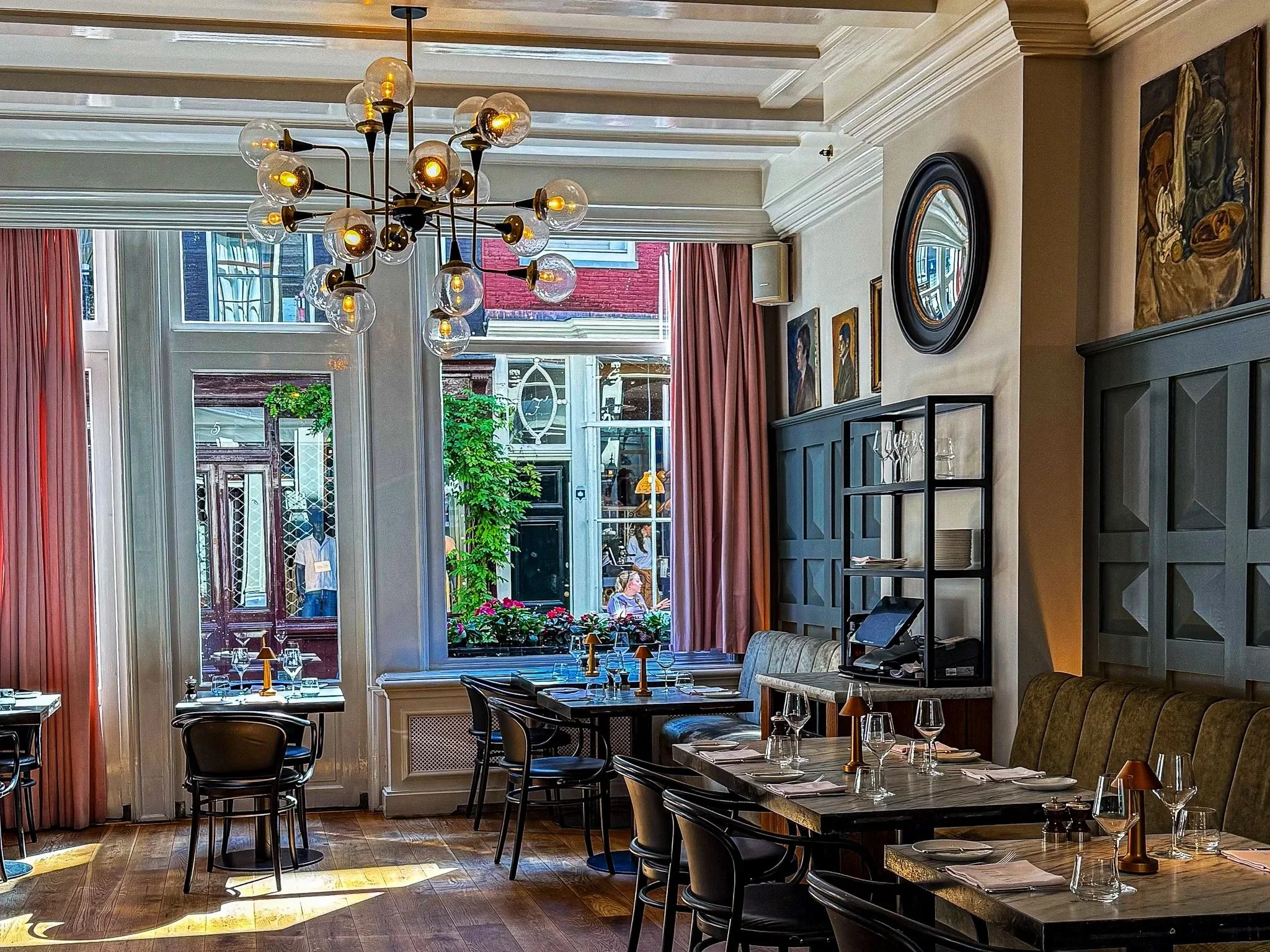 Interior of a restaurant with wooden tables set with wine glasses, napkins, and small lamps. Large windows with pink curtains and a modern chandelier overhead.
