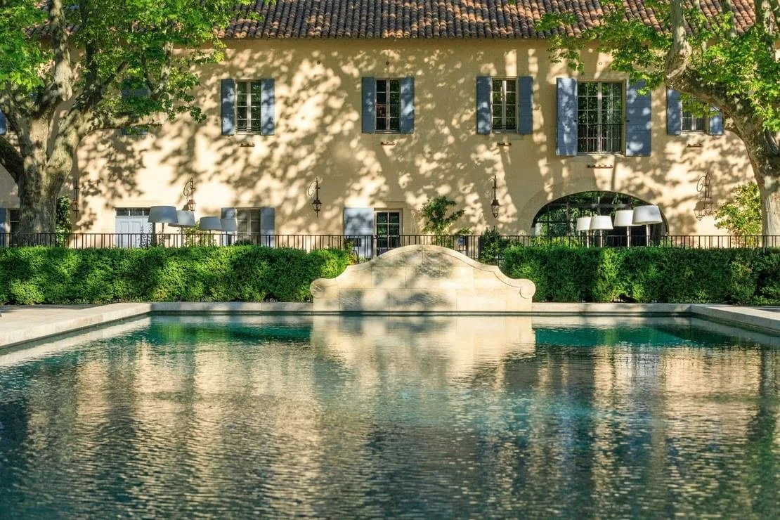 Reflected swimming pool in front of a yellow stone Provençal estate with pale blue window shutters shaded by large plane trees.