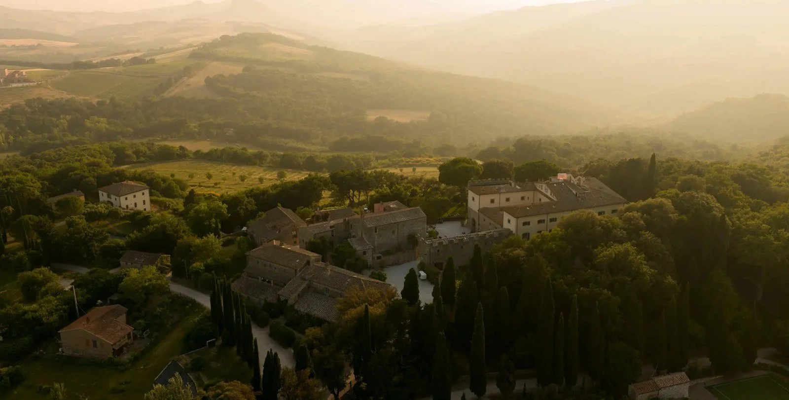Aerial view of Borgo Pignano estate in Tuscany at golden hour, stone hamlet buildings ringed by cypress trees, woodland and hills.