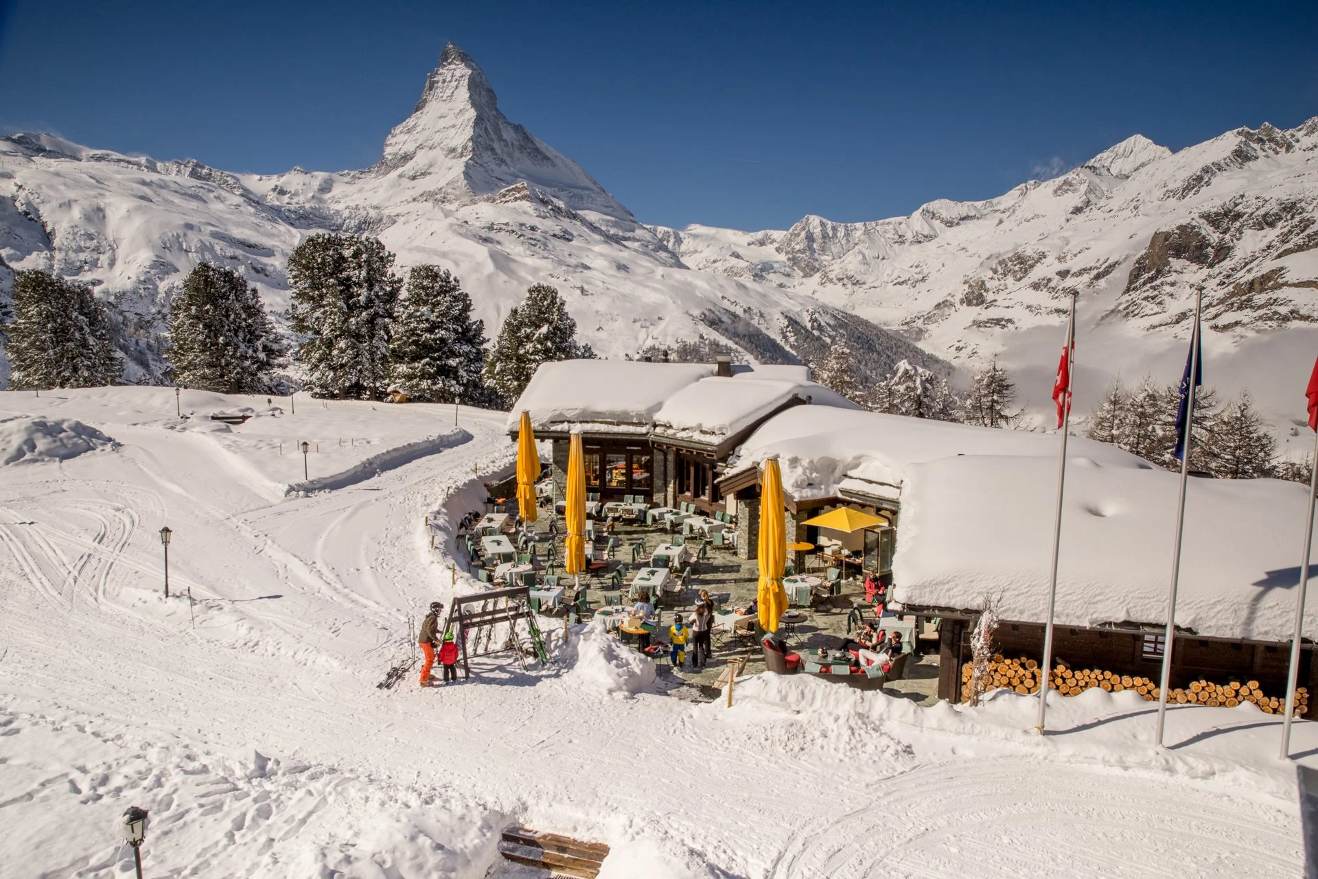 A snowy mountain landscape with a chalet and outdoor seating area, surrounded by snow-covered trees, with Matterhorn mountain in the background.