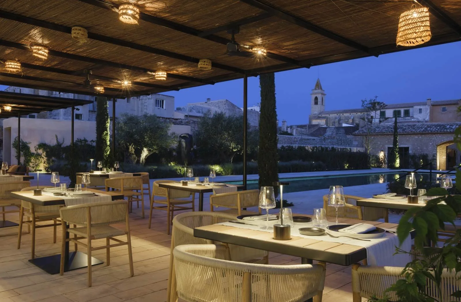 Poolside dining terrace at Can Ferrereta in Santanyi, Mallorca, woven chairs under a reed pergola with the village church tower beyond at dusk.