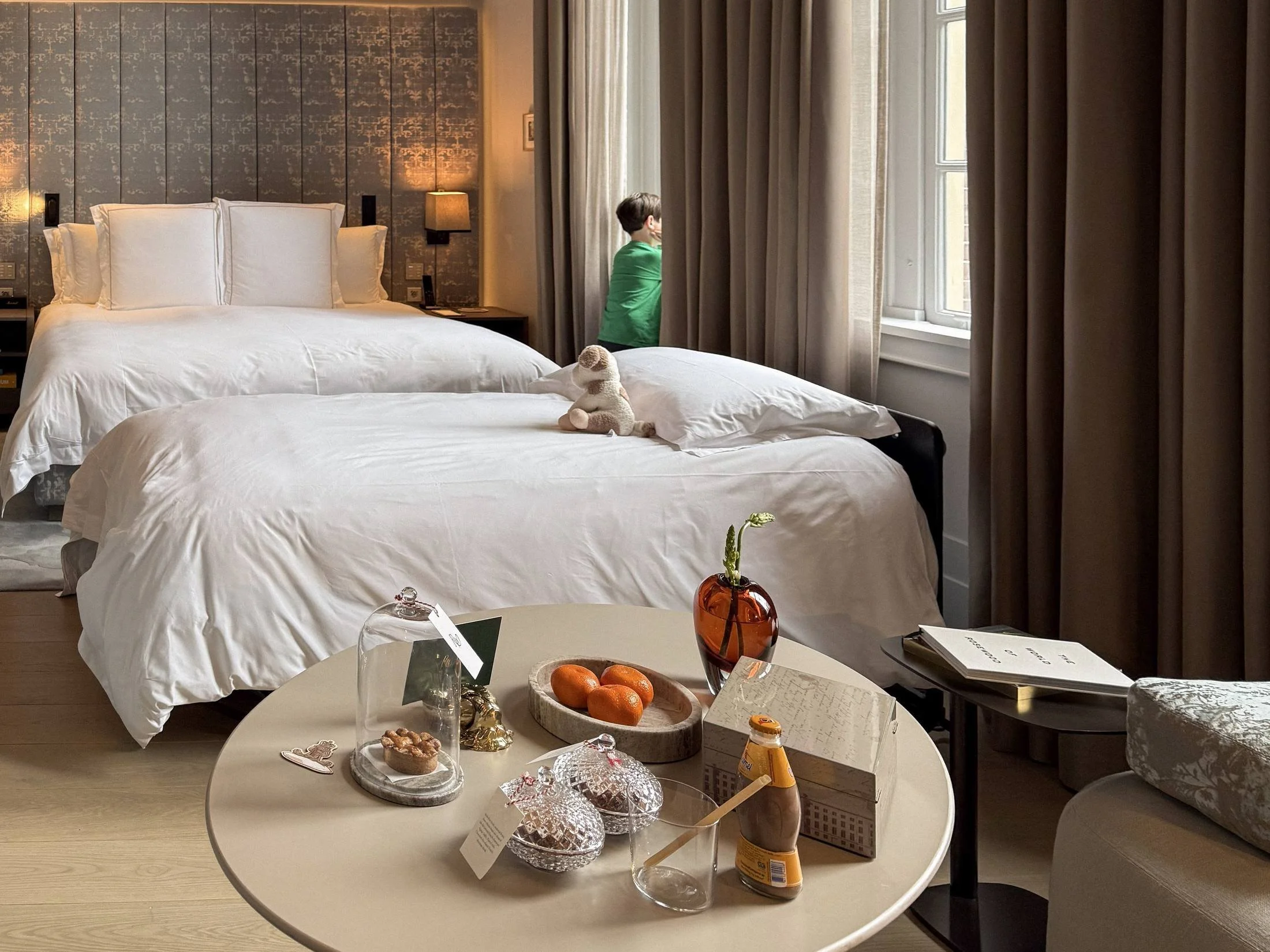 Guest room at Rosewood Amsterdam with twin beds, a stuffed animal, tall curtained windows and a welcome amenity tray on a round table