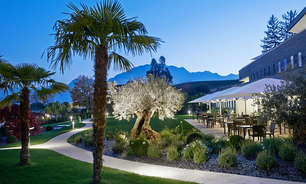 Outdoor garden with palm and flowering trees, a winding path, outdoor dining area with umbrellas, mountains in the background, and evening lighting.