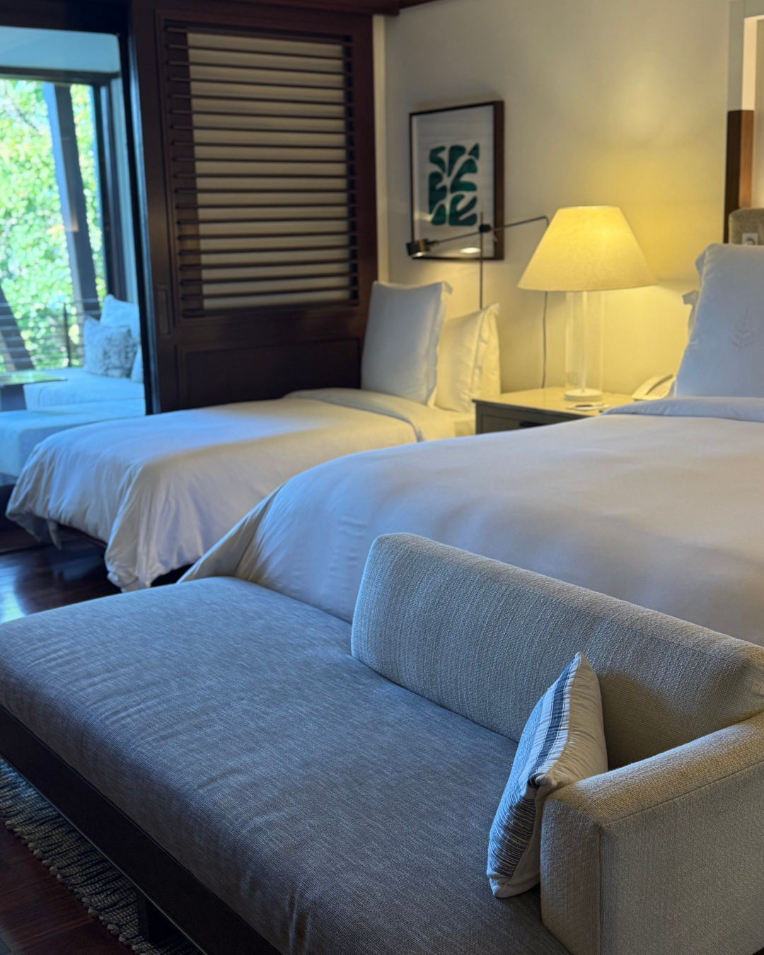 Hotel room with two neatly made beds, a lamp on a nightstand, artwork on the wall, and a seating area with outdoor view.