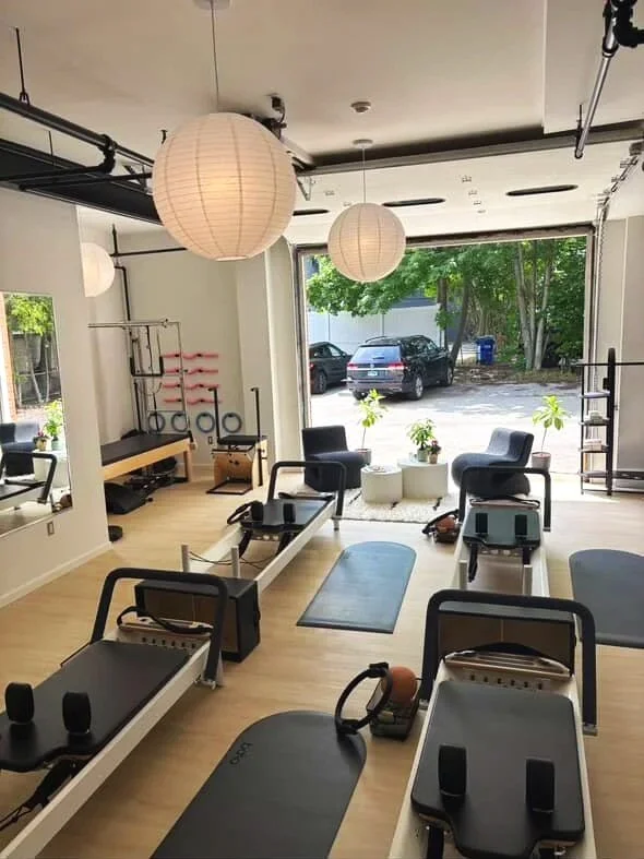 Interior view of a Pilates or fitness studio with exercise equipment, large window showing outdoor trees and parked cars, and hanging paper lantern lights.