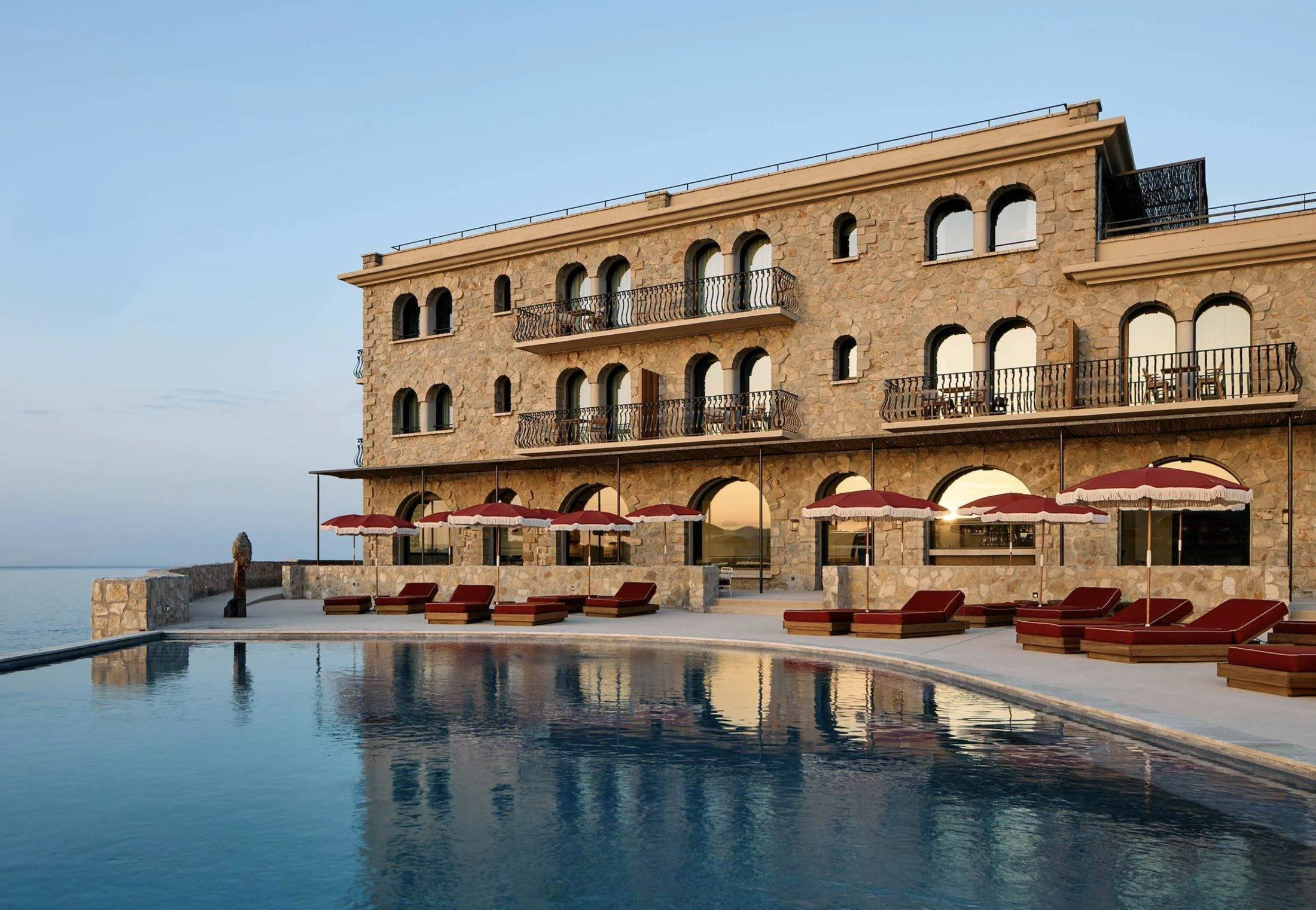 Pool, red fringed umbrellas and arched stone facade at dusk at Zannier Île de Bendor, the private island resort off Bandol on the French Riviera