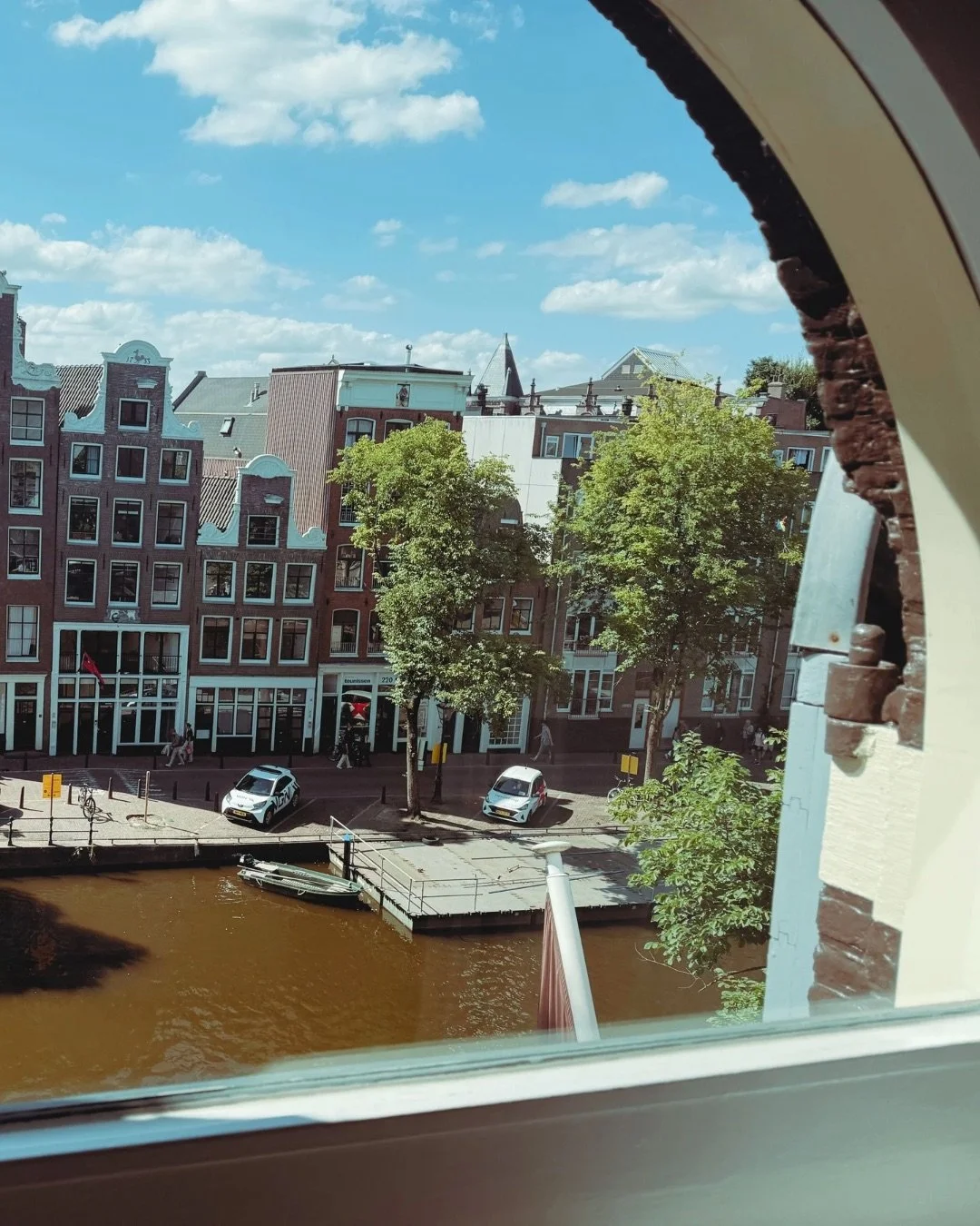 View through a circular window showing a canal with brown water, trees, parked cars, and historic buildings in a city, under a blue sky with scattered clouds.
