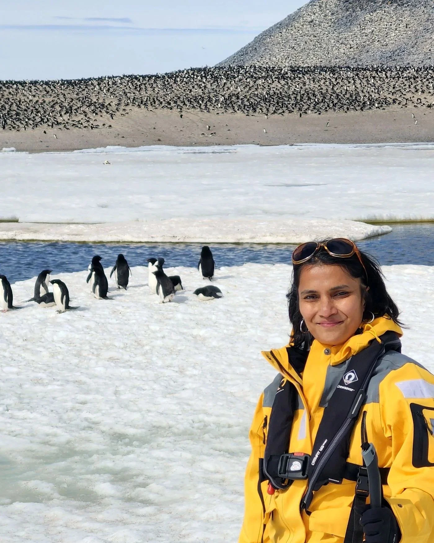 Expert Travel Advisor Dee Swaminathan in a yellow jacket on the ice in antarctica with a penguin colony in the background