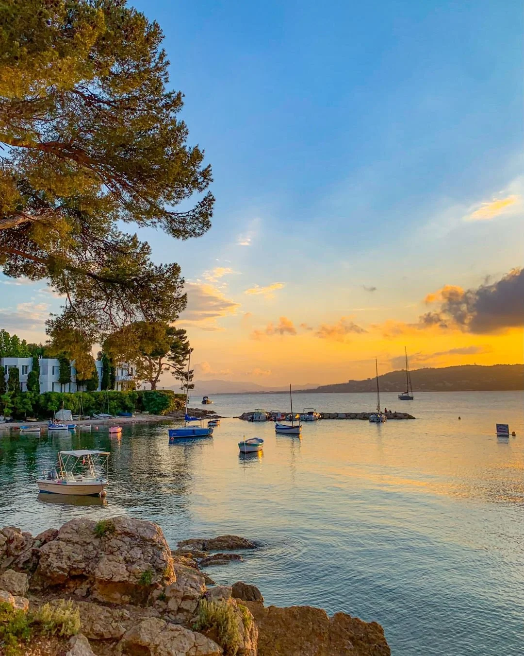 Scenic view of a harbor during sunset with small boats anchored in the water, a rocky shoreline in the foreground, and colorful clouds in the sky.