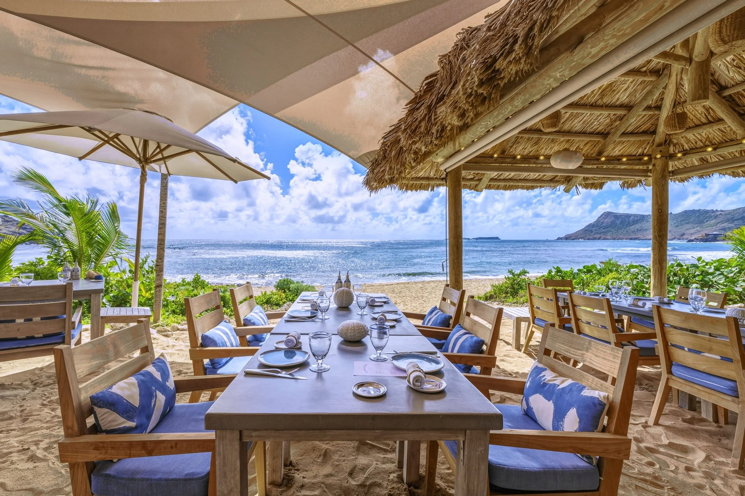 Beach club dining at Hotel Le Toiny in St. Barth, a long table under a sailcloth canopy and thatched roof facing the sea and a headland.
