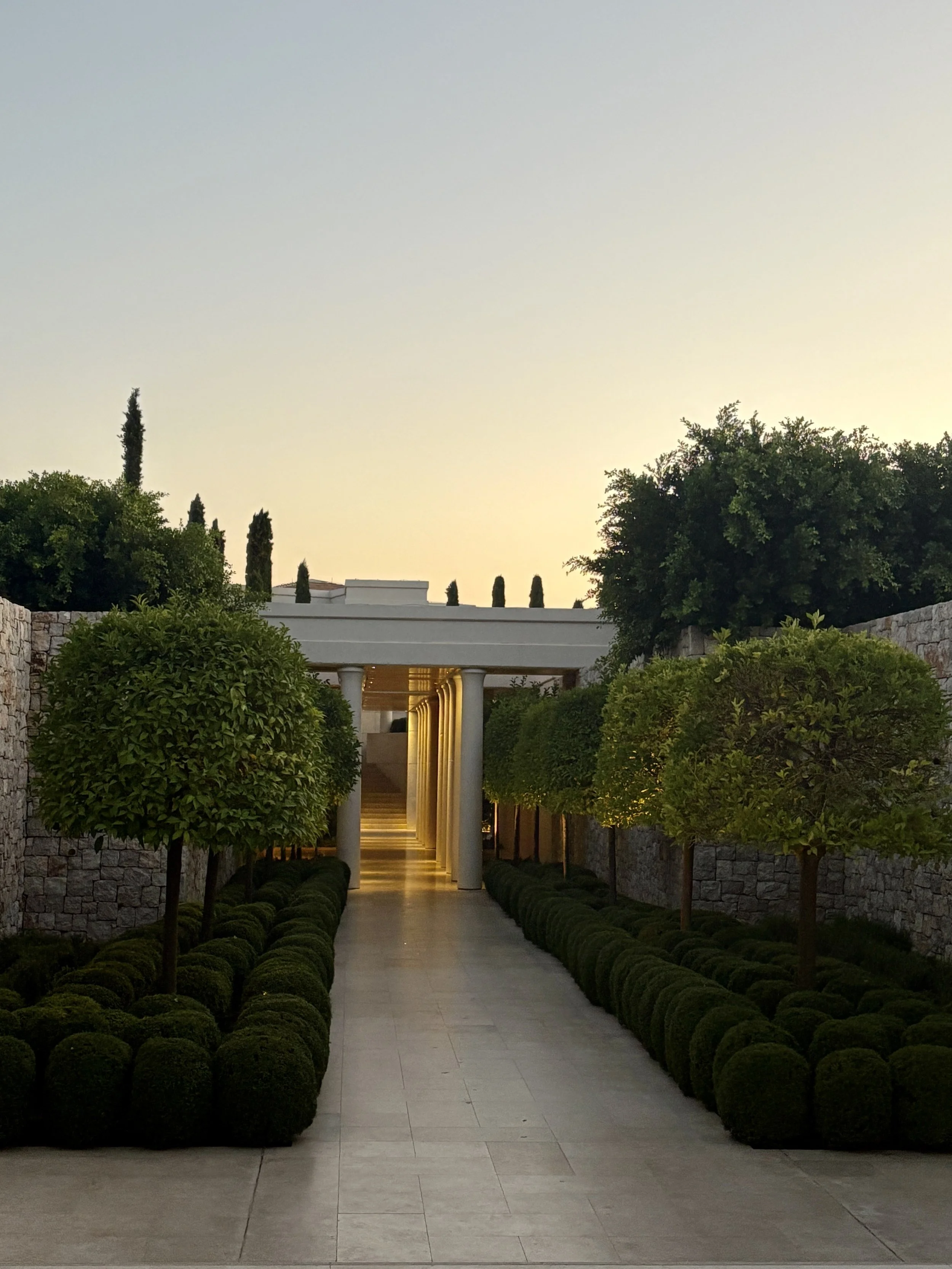 Symmetrical garden pathway lined with trimmed bushes and trees, leading to a classical white colonnade under a clear sky at sunset.