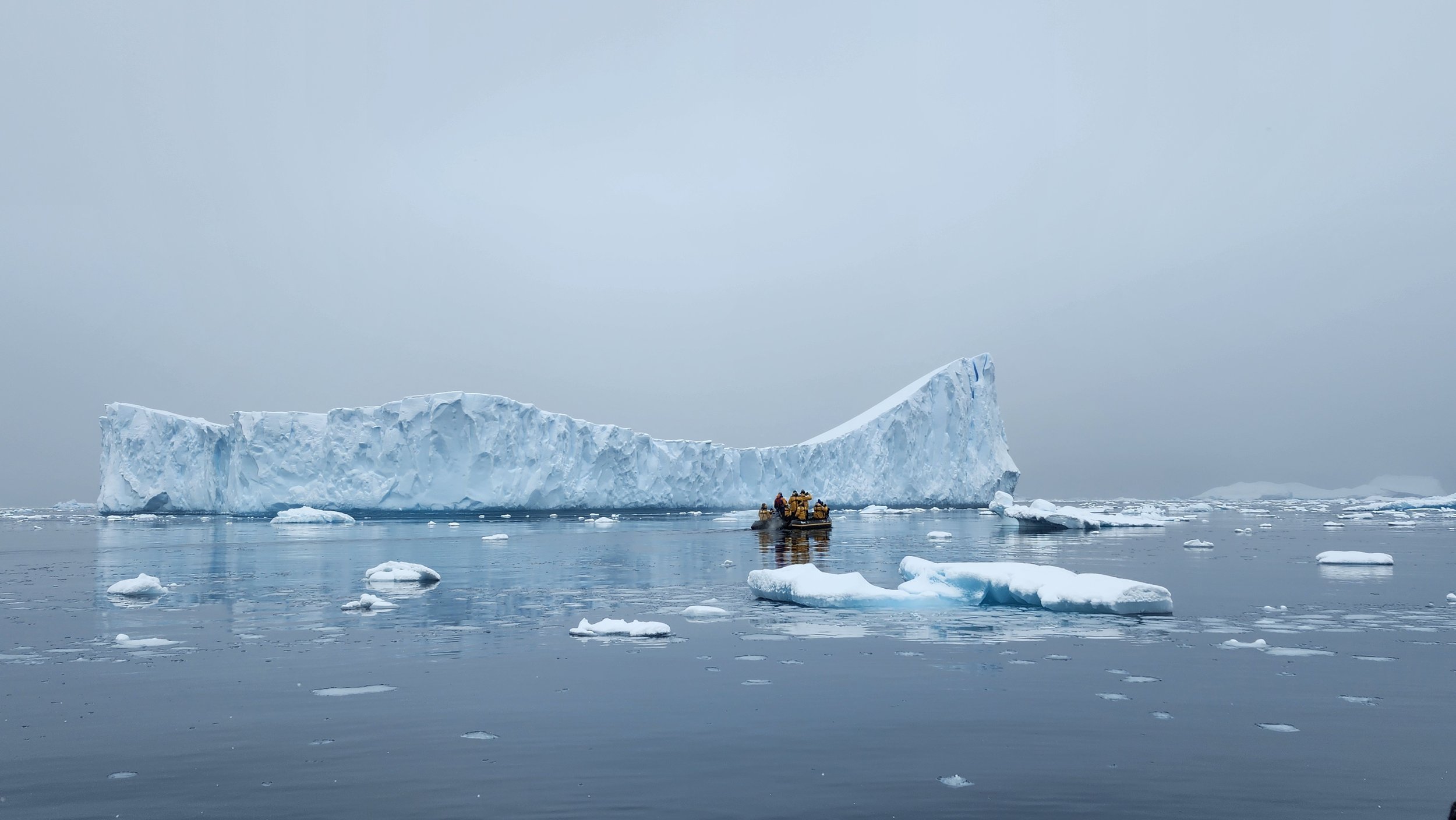 A small group in yellow expedition parkas rides a Zodiac past a large blue-white iceberg in calm, ice-strewn Antarctic water under a flat gray sky.