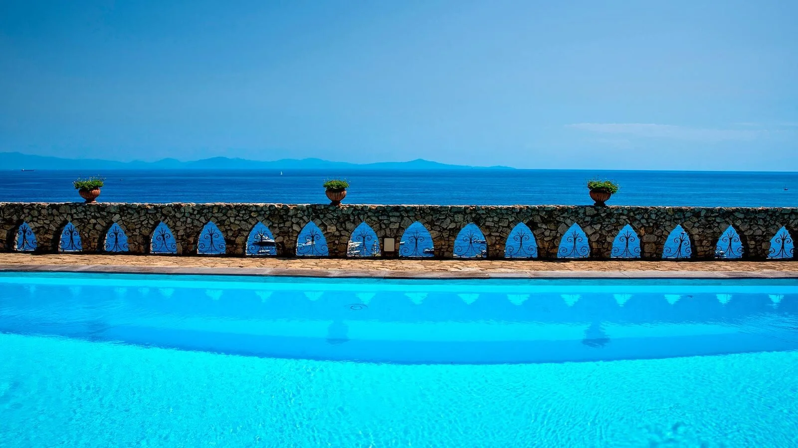 Piscina exterior de agua salada en Borgo Santandrea, con un muro de piedra con aberturas arqueadas de color azul que enmarcan la costa amalfitana