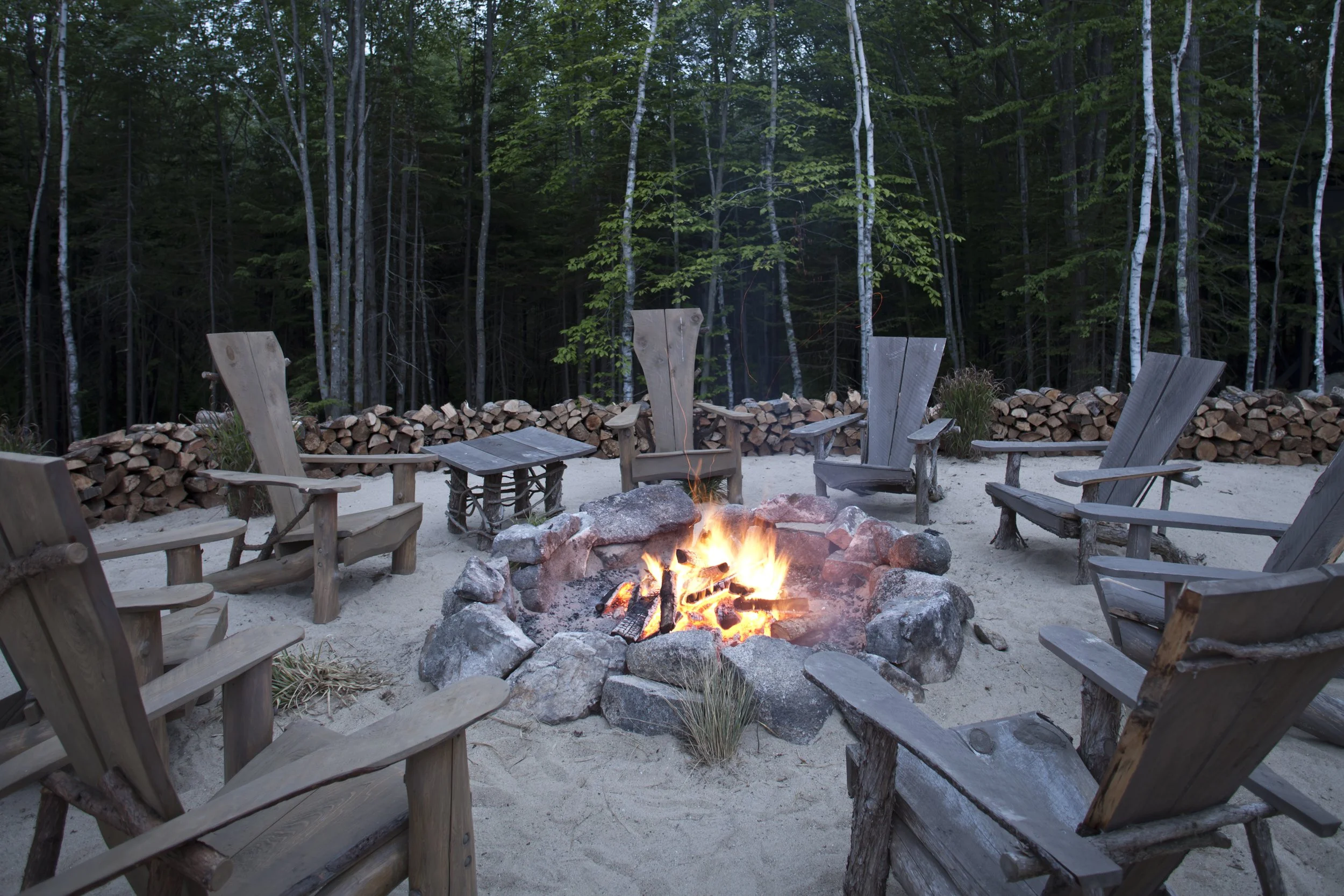 A circle of wooden Adirondack chairs surrounds a campfire on sand with a backdrop of densely wooded trees and stacked firewood.