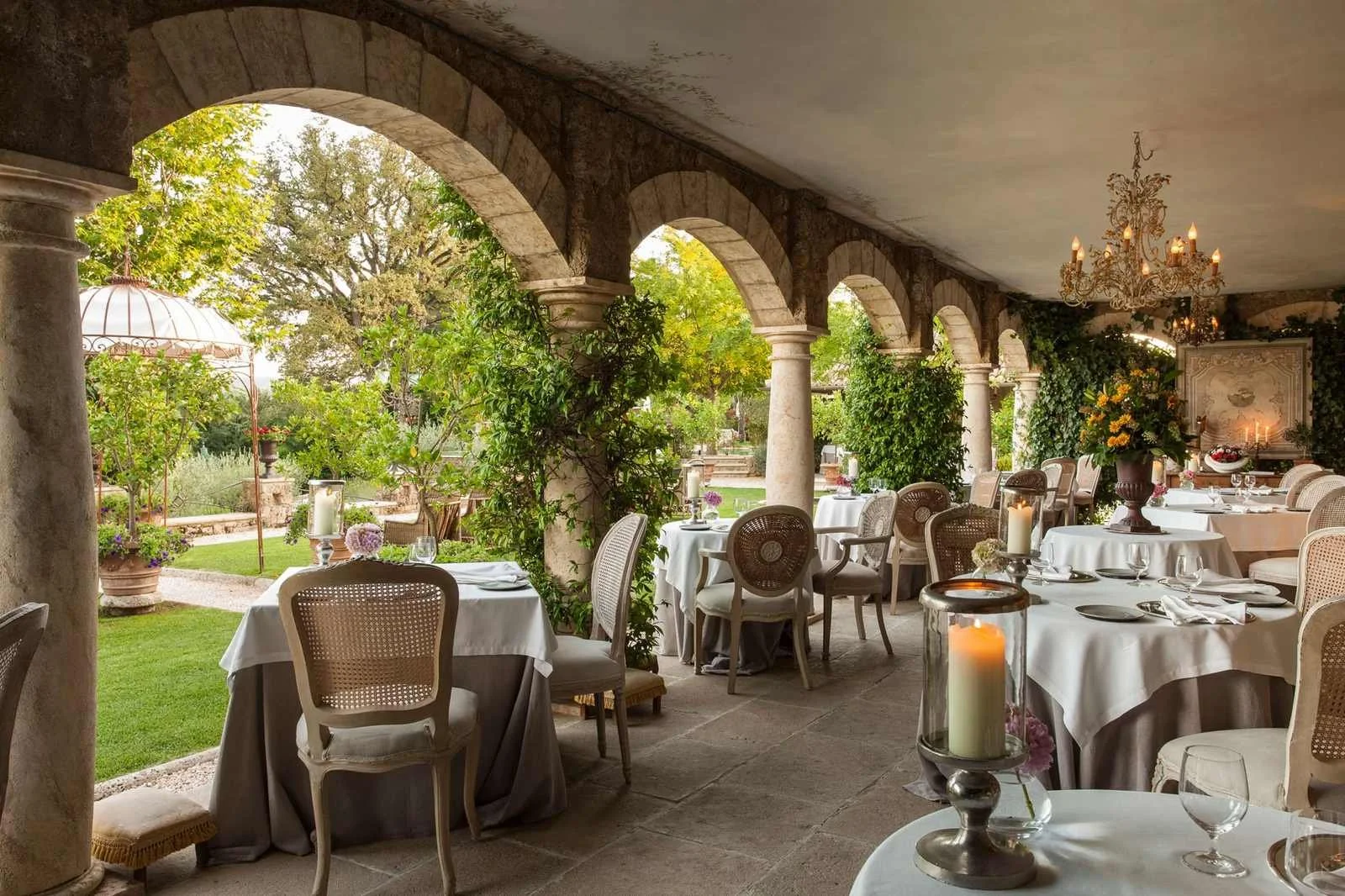Stone arched garden loggia set for dining at Borgo Santo Pietro in Tuscany, white-linen tables and cane chairs under a chandelier, gardens beyond.