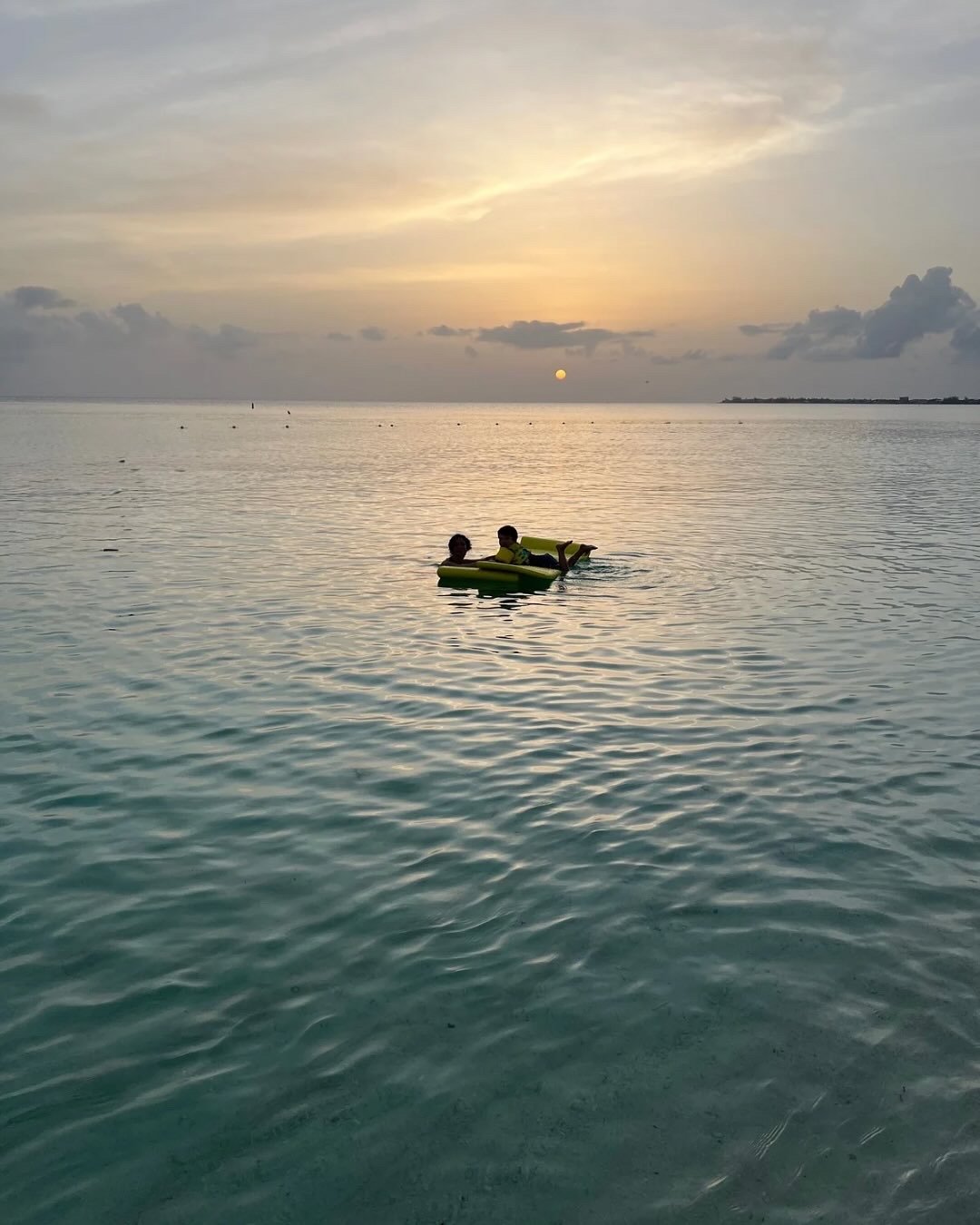 日落时分，两个孩子躺在平静水域的浮筏上，背景是云朵与远处的海岸线。
