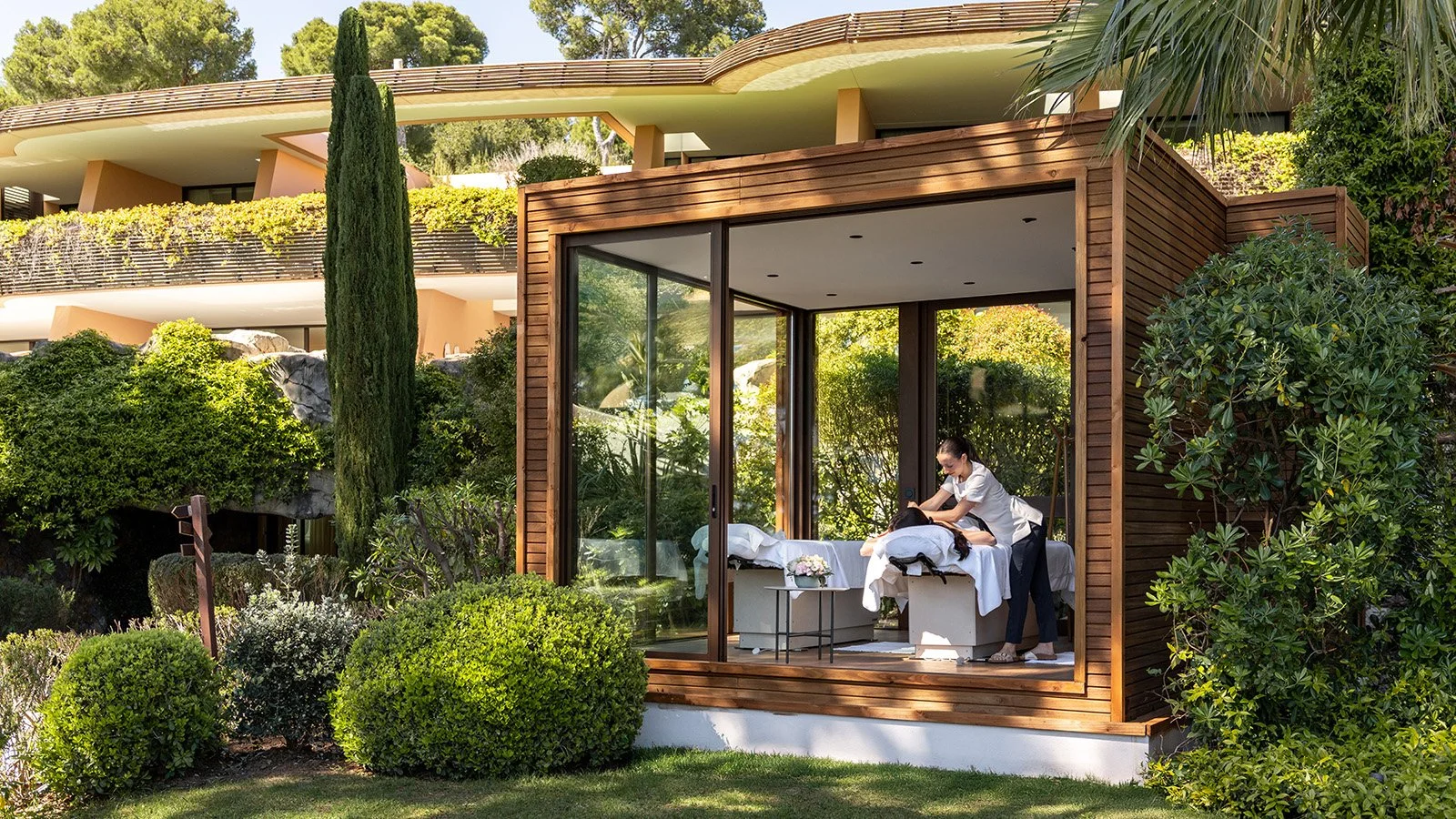 Modern wooden and glass outdoor spa cabana surrounded by lush greenery, featuring a massage table and therapist inside.