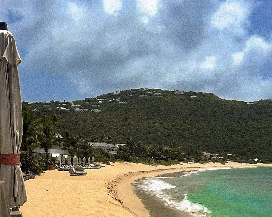 The wide sweep of Flamands Beach at Cheval Blanc St-Barth, with loungers under the palms, surf breaking along the shore and villas scattered across the hillside above the bay.