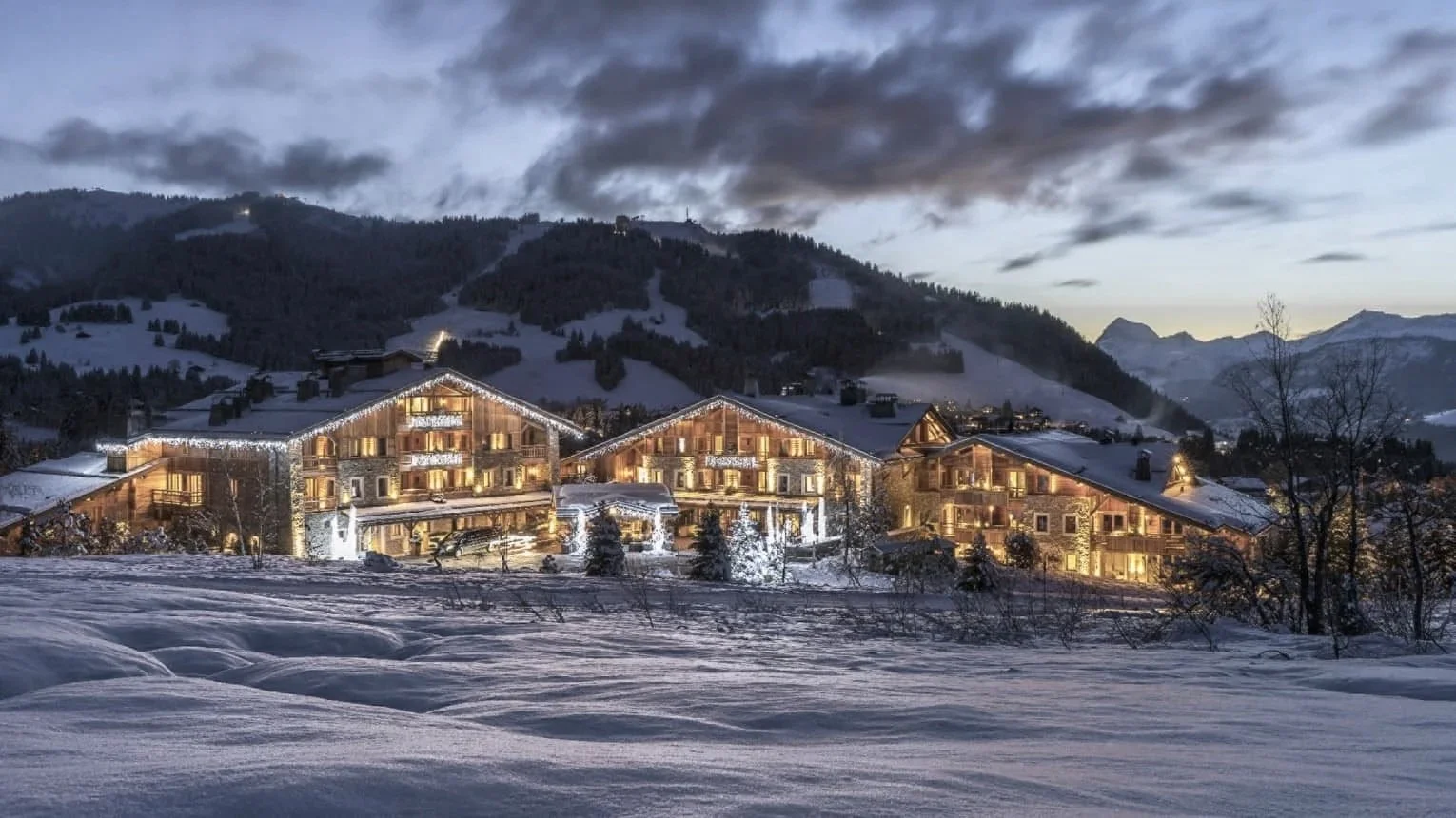 Lit alpine chalets at Four Seasons Hotel Megeve at dusk, with the snow-covered slopes of Mont d'Arbois behind