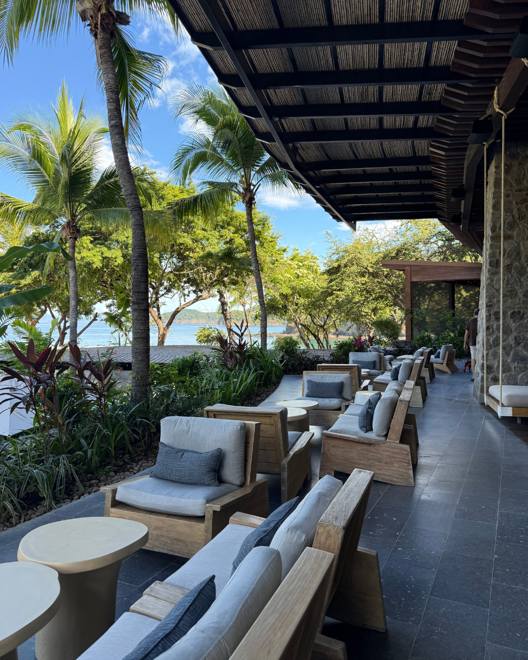 户外休憩区配备木质座椅与靠垫，白天可俯瞰热带景观：棕榈树、绿植与背景中的水域相映成趣。