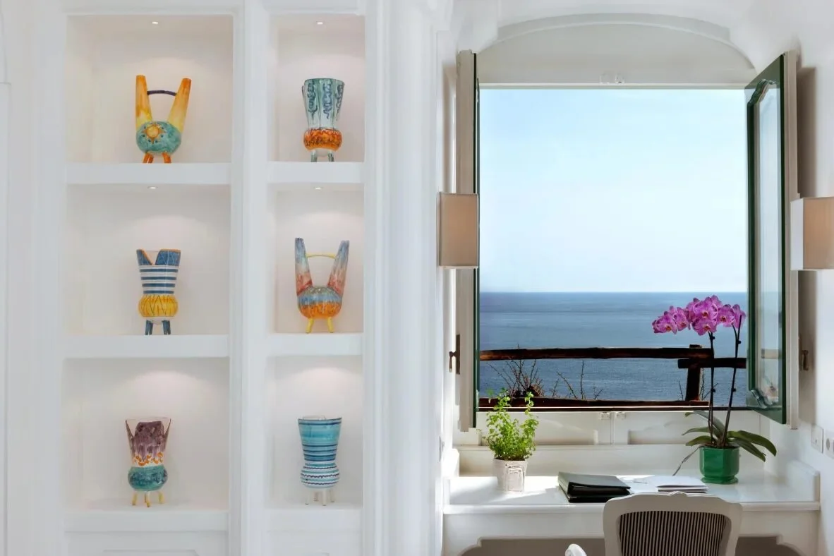 Cerámica de Vietri en estanterías iluminadas junto a una ventana abierta con vistas al mar en Il San Pietro di Positano
