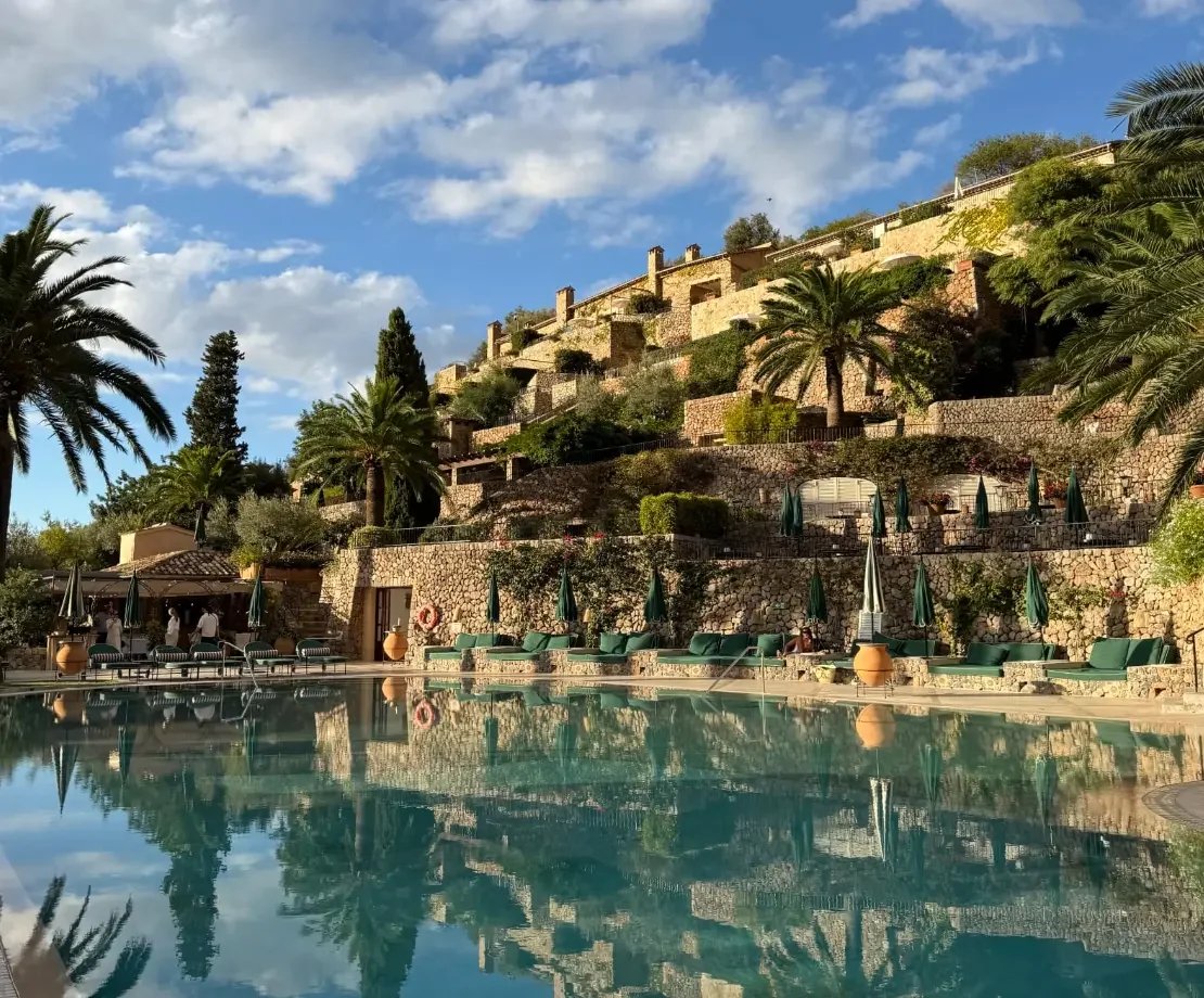 Outdoor swimming pool surrounded by terraced stone buildings, palm trees and loungers at La Residencia in Deià, Mallorca.